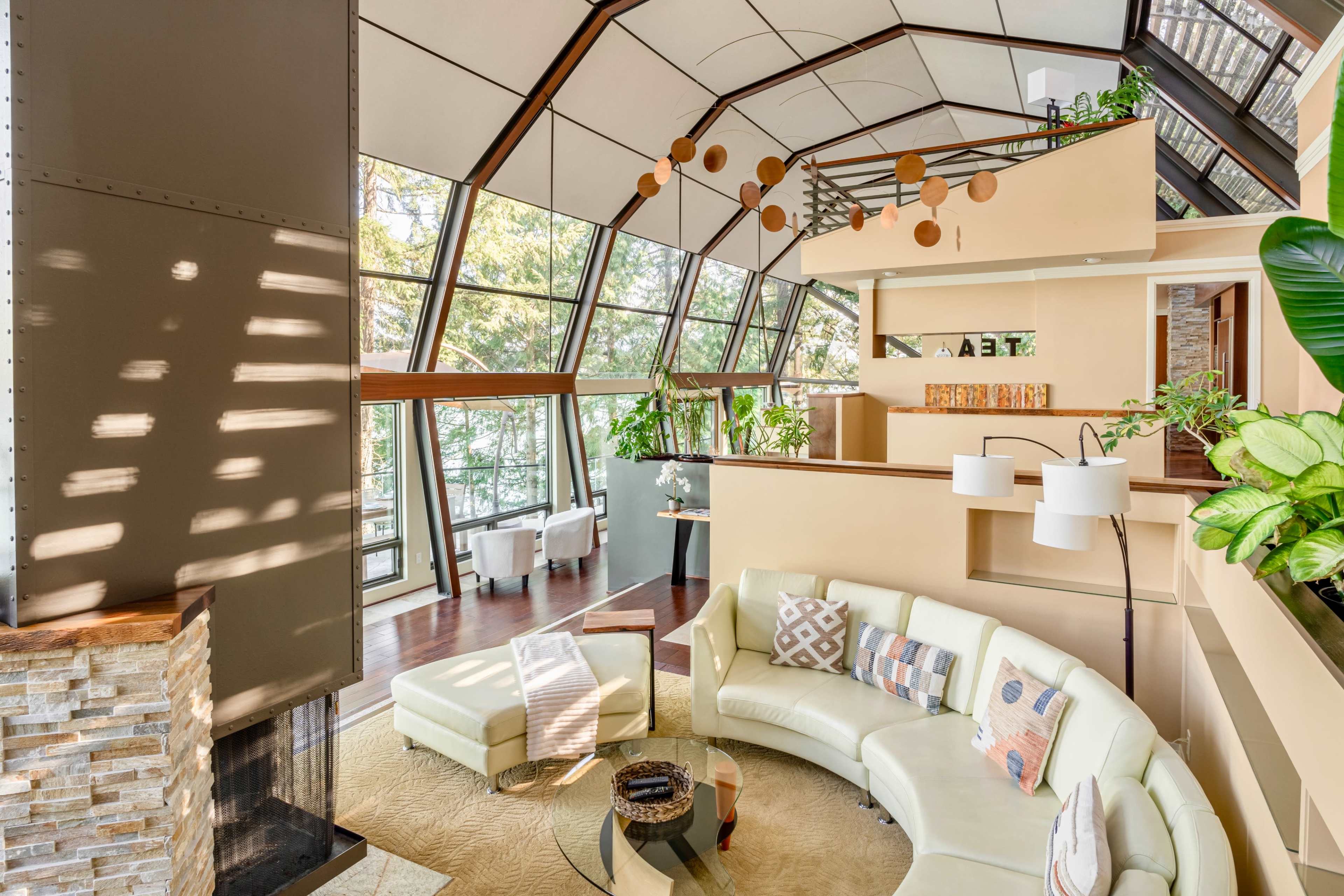 A spacious, modern living room with large windows, a curved white sofa, and various plants, creating a bright and airy atmosphere.