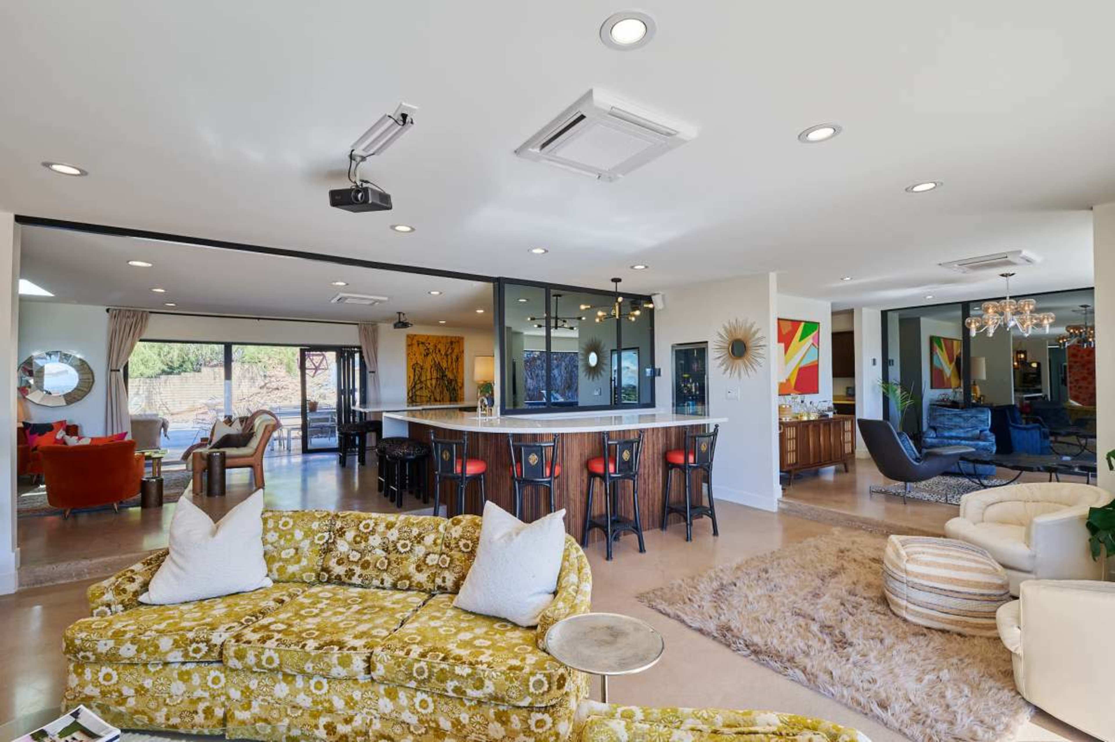 The open-concept living area features a mix of colorful furniture, a modern kitchen bar, and large windows overlooking a patio.