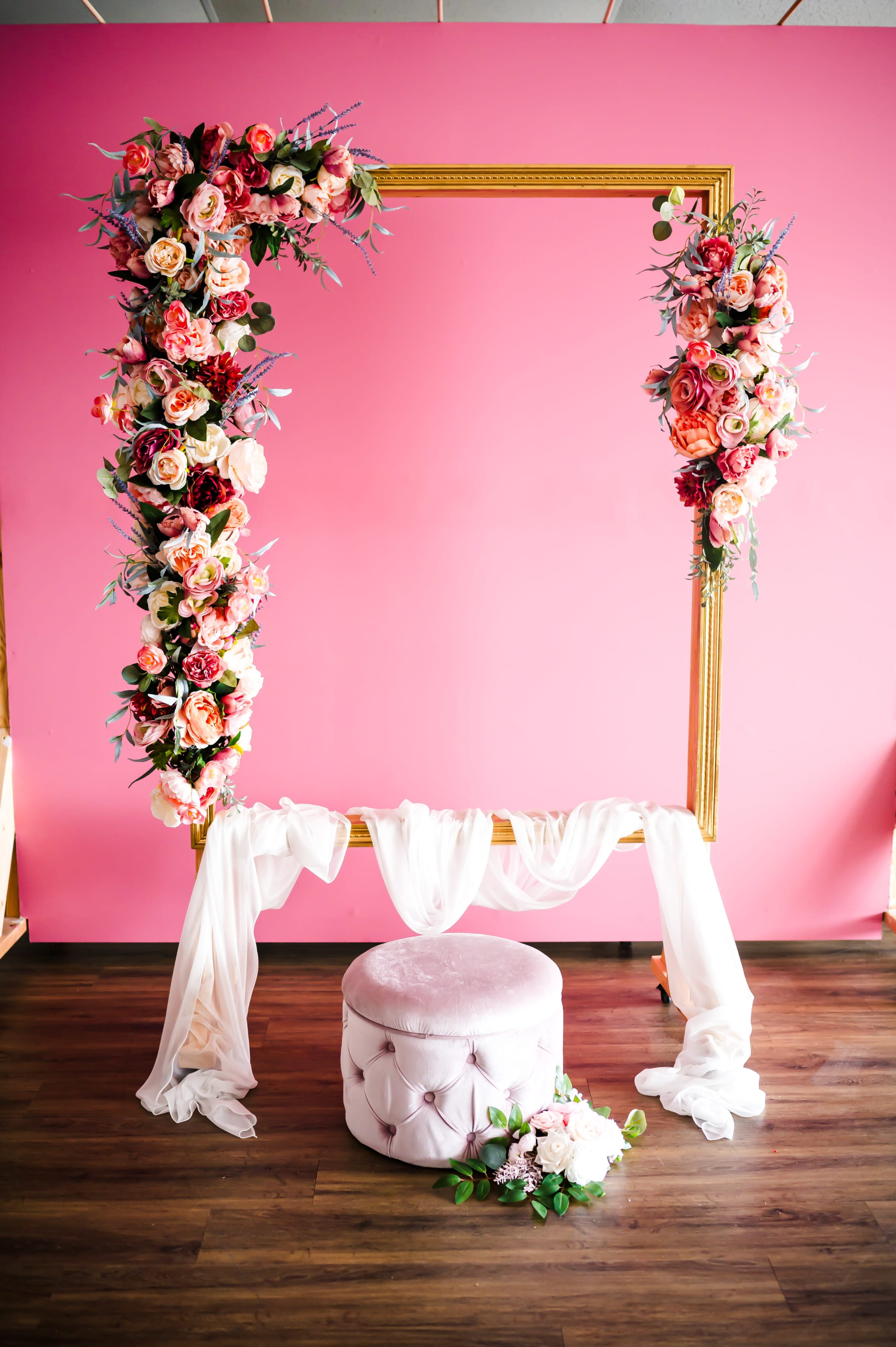 A large ornate frame adorned with flowers is positioned against a pink wall, with a plush stool and draped fabric beneath it.