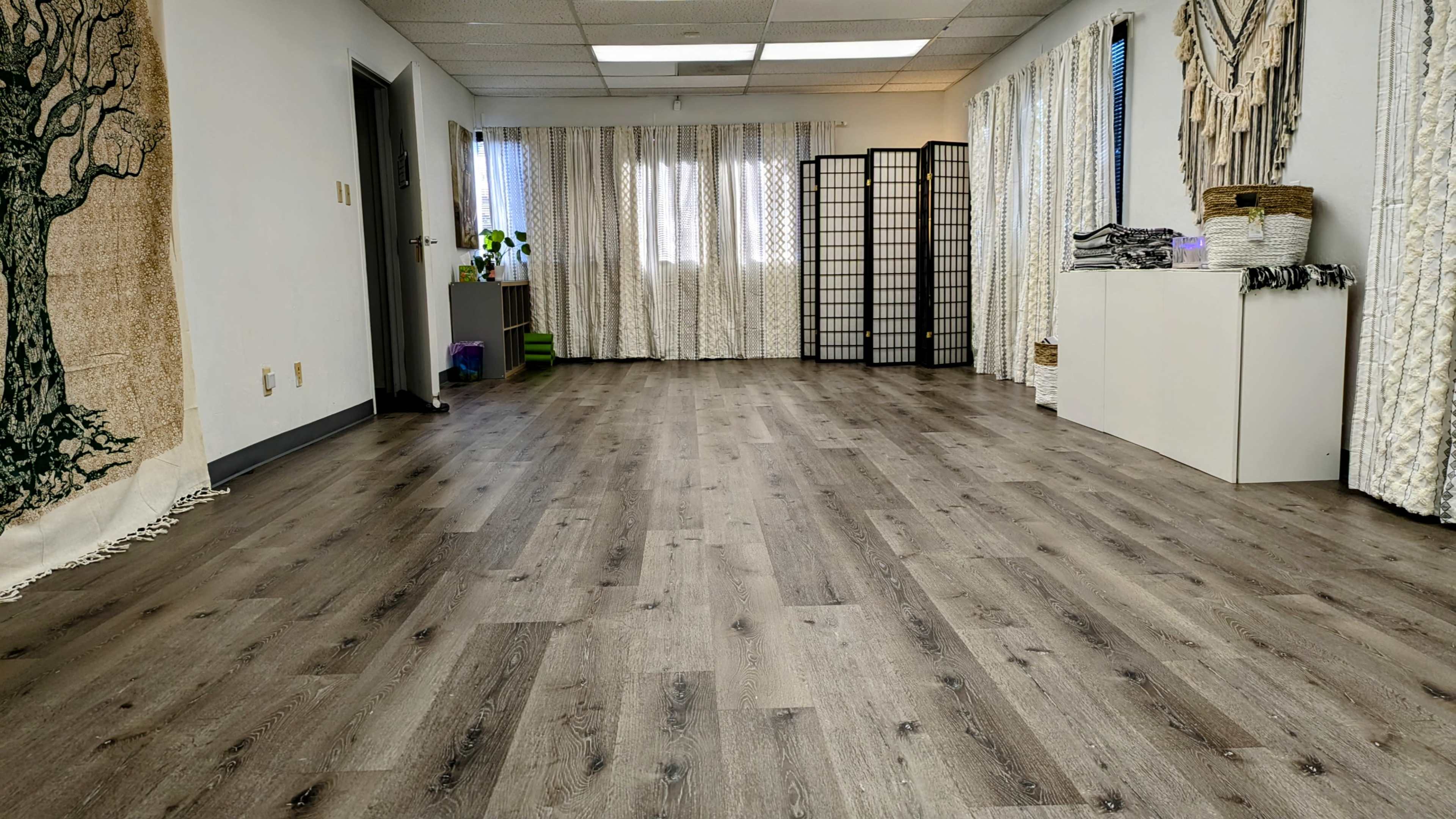 The image shows a spacious, well-lit room with wooden flooring, light-colored walls, and decorative elements such as a tapestry and folding partitions.