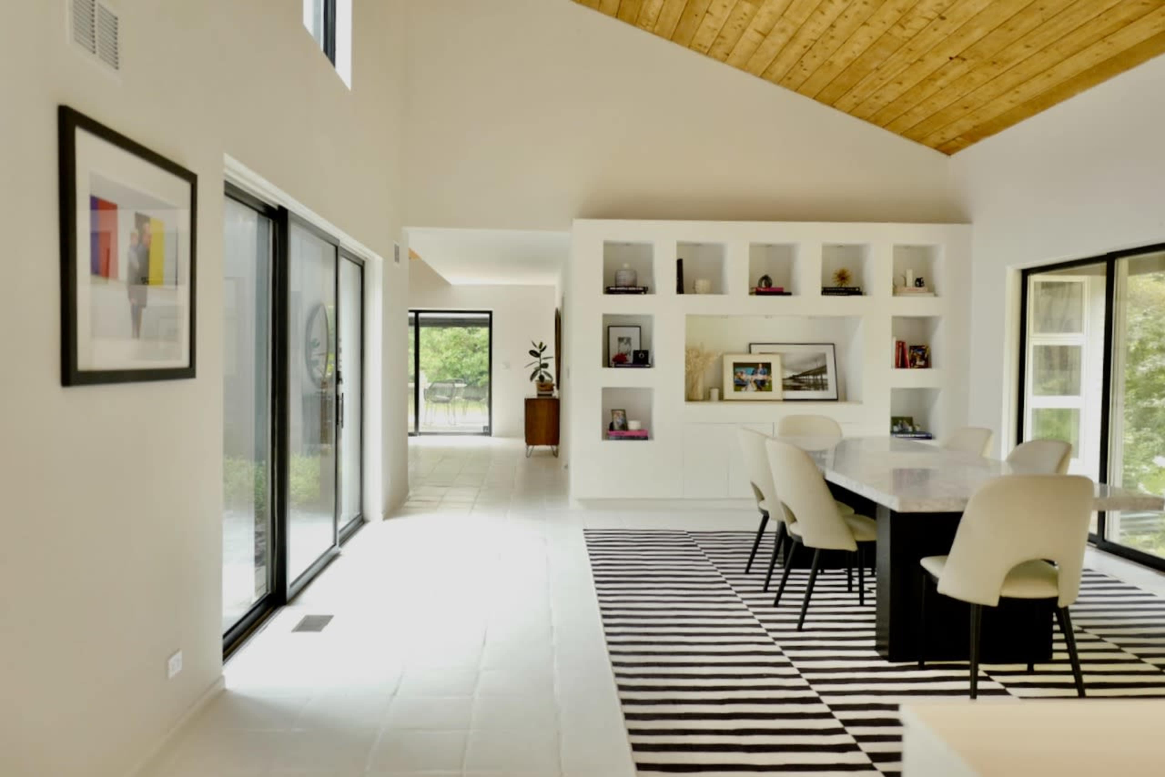 A spacious dining area with a large table surrounded by chairs, framed by large windows and a wall of shelves, featuring a modern wooden ceiling.