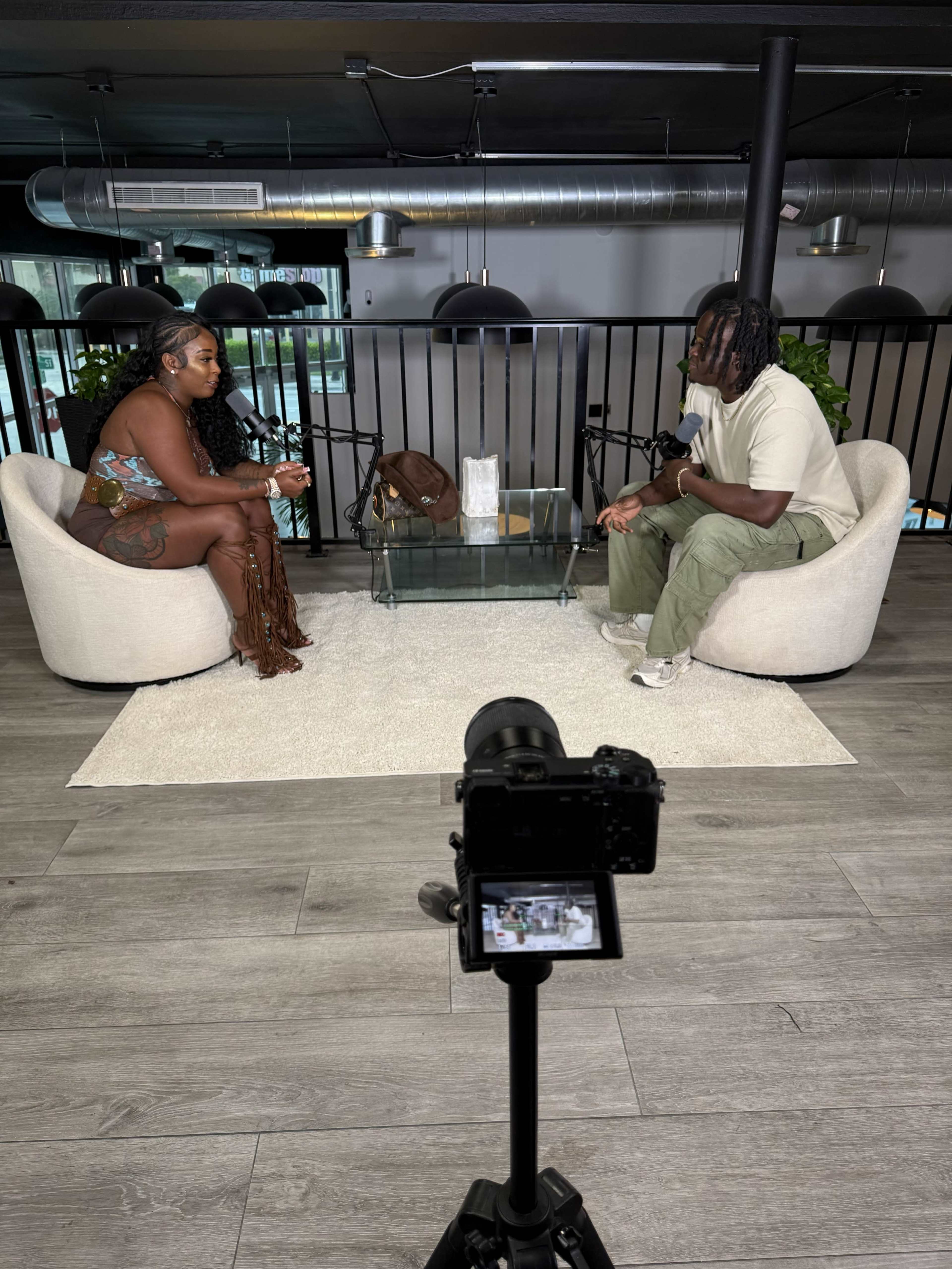 Two individuals are seated in modern chairs across from each other in a well-lit studio, with a camera on a tripod focused on their conversation.