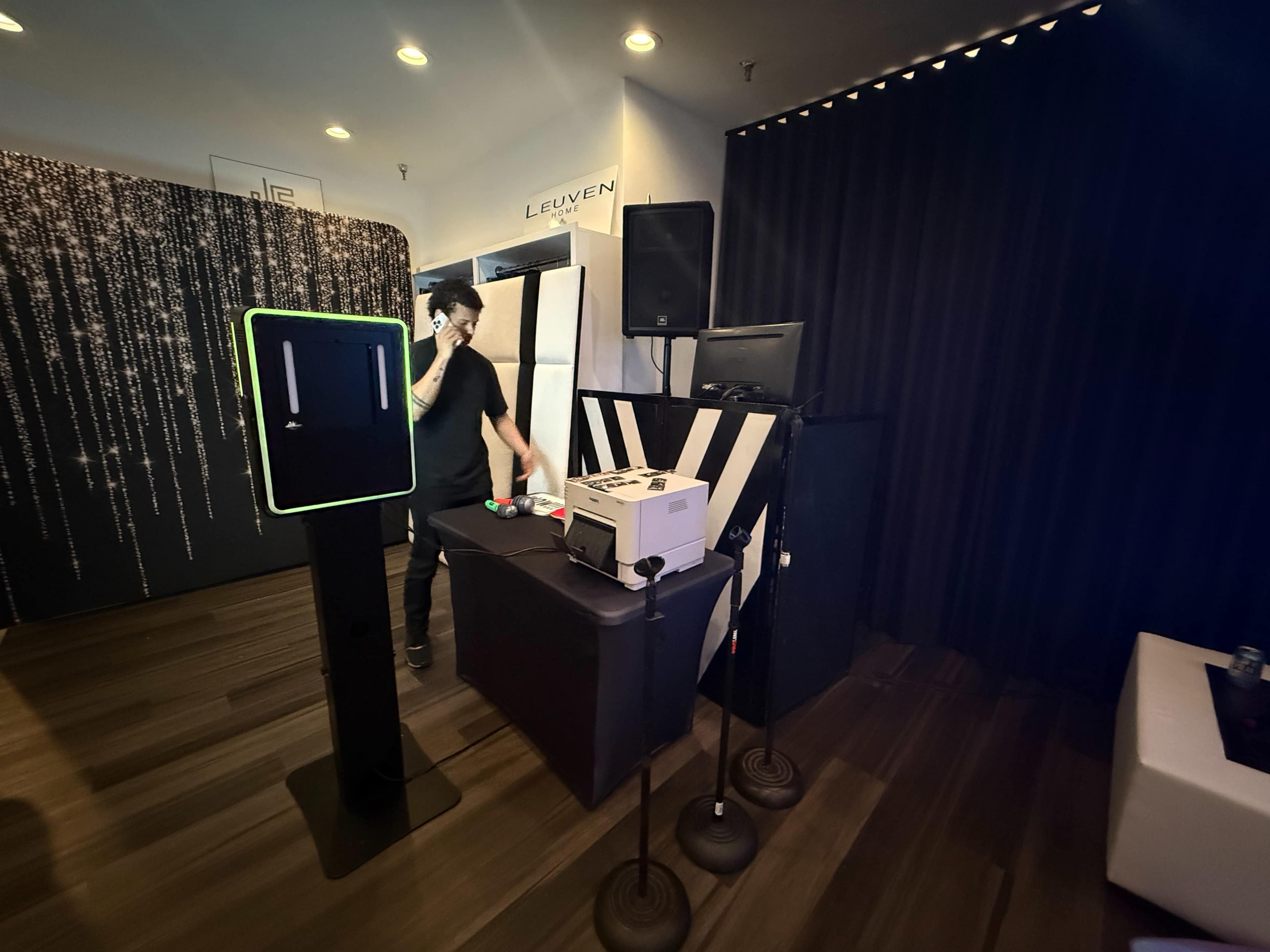 A person stands behind a table with a white box and electronic equipment in a dimly lit room decorated with sparkling curtain drapes.