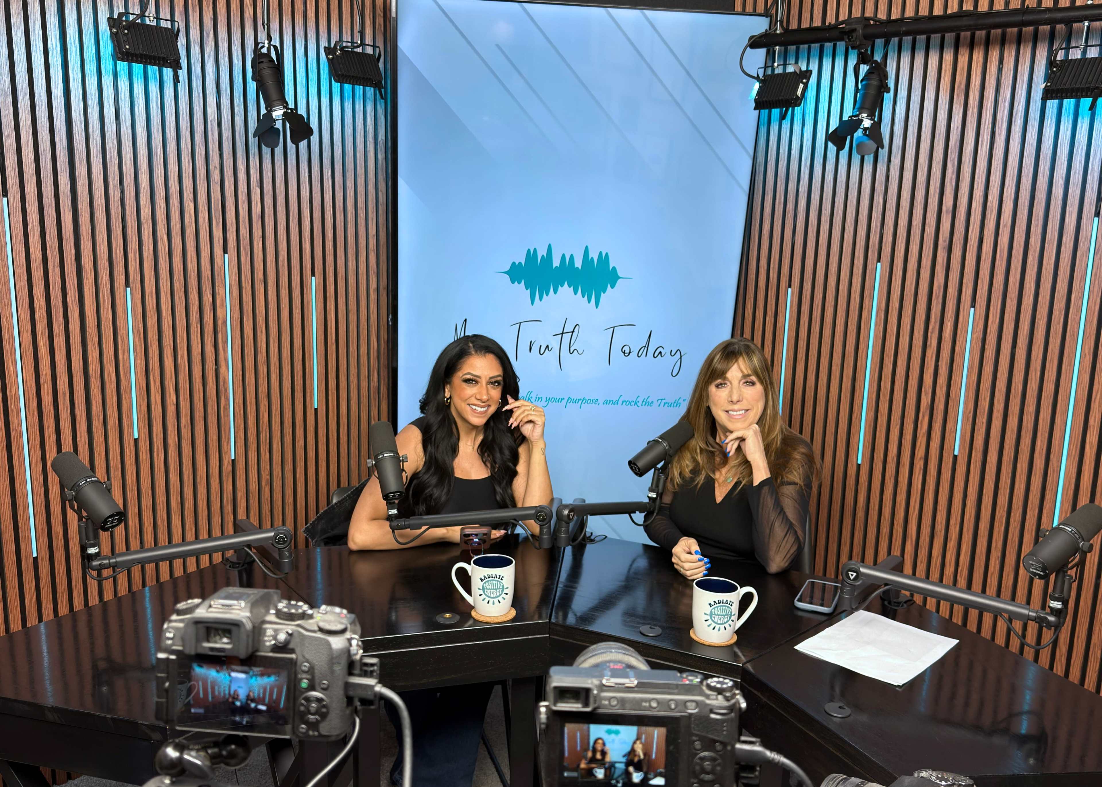 Two women are sitting at a table in a studio, smiling and facing the camera, with microphones in front of them and a backdrop that reads "New Truth Today."