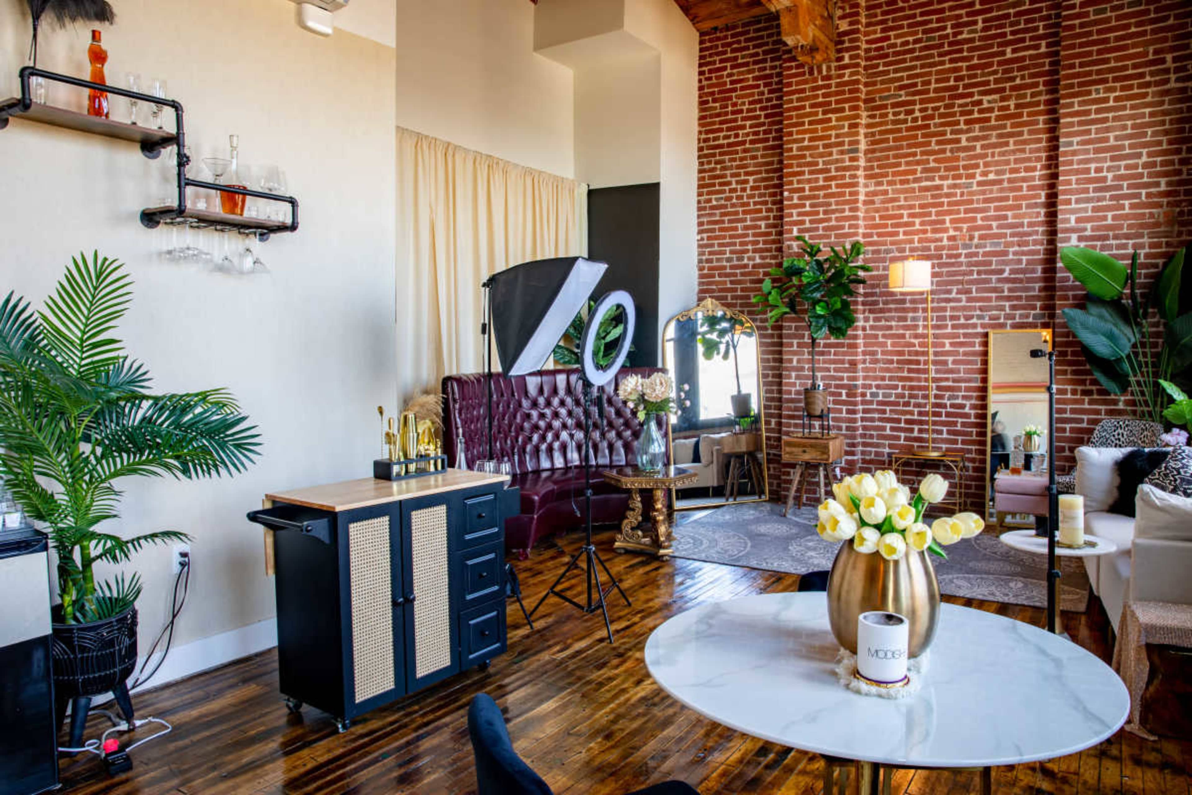 A stylish interior setting featuring a mix of brick walls, a tufted burgundy sofa, a round marble table with a vase of tulips, and various decorative plants.