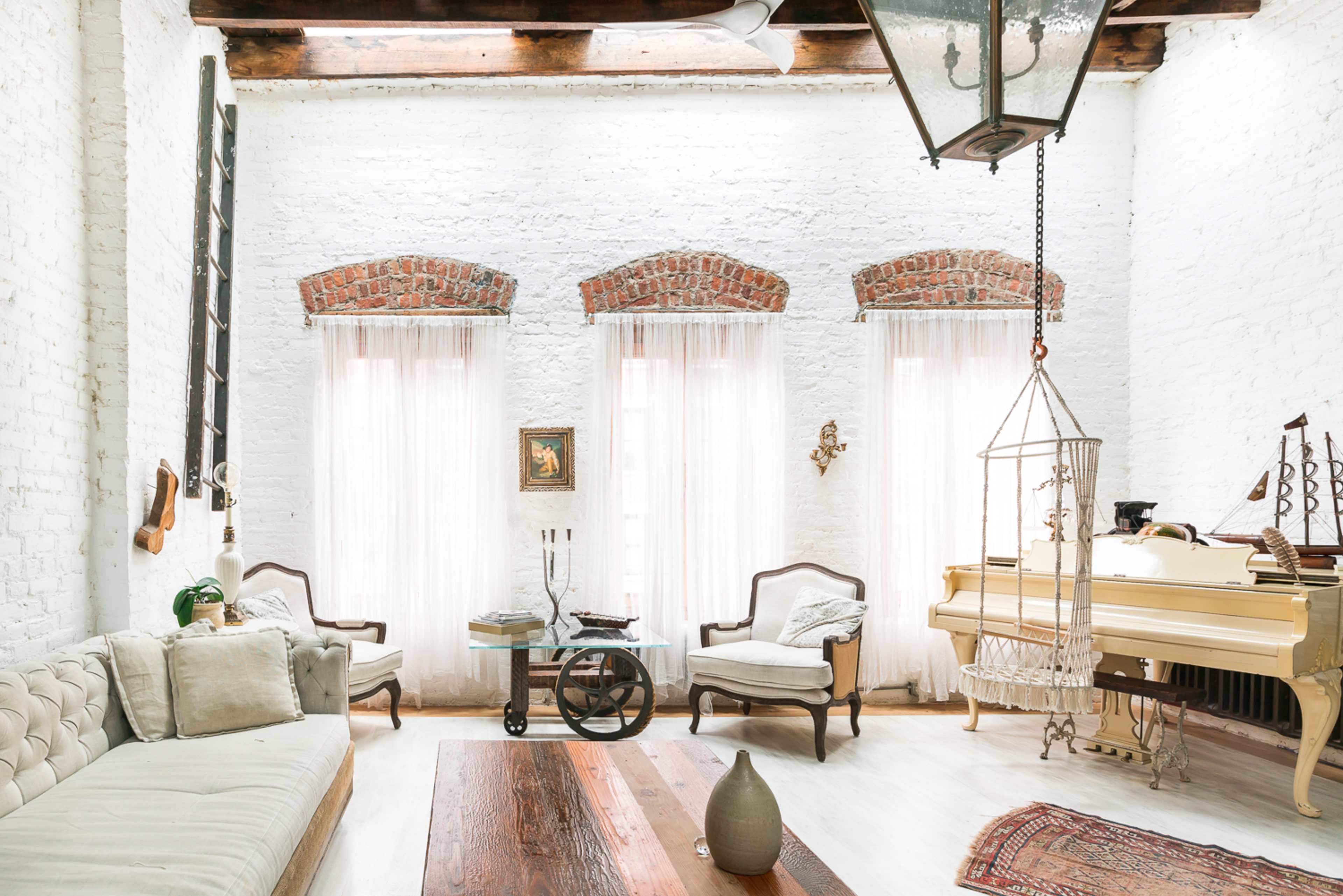 A bright, airy living room with white brick walls, large windows draped in sheer curtains, a grand piano, and vintage furniture arranged around a wooden coffee table.