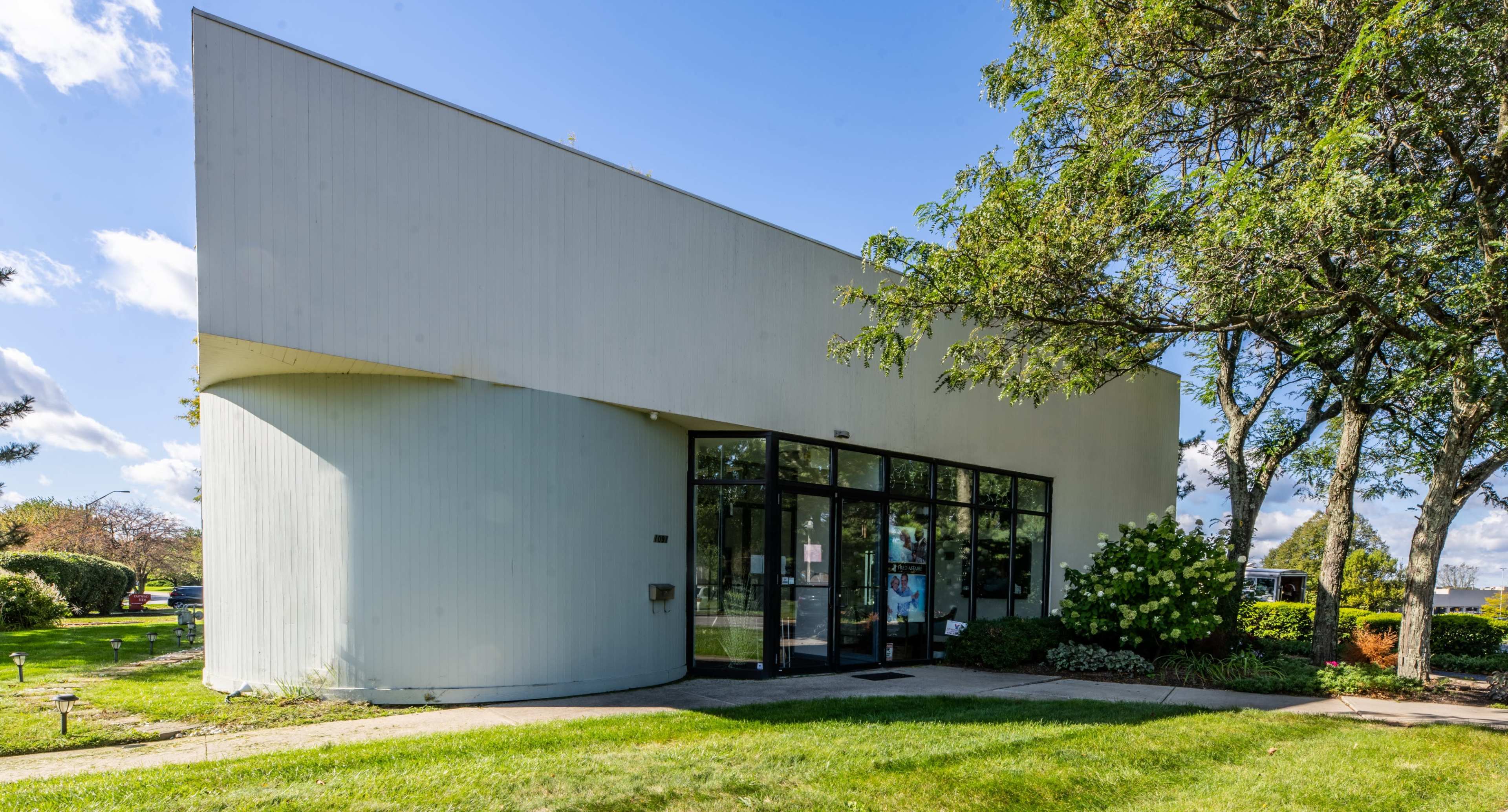A modern, curved white building with large glass doors and windows, surrounded by well-maintained greenery and trees.