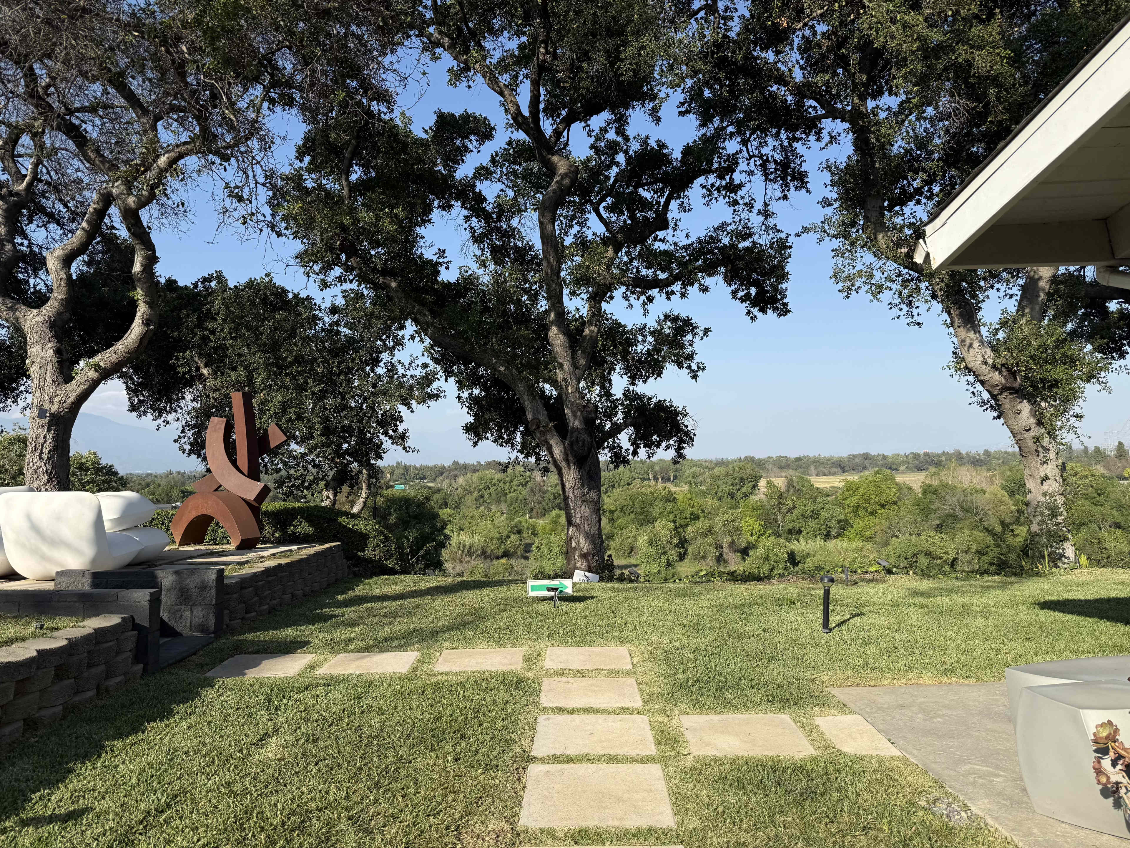 The image shows a landscaped outdoor area with stone pavers, a sculpture, and lush trees overlooking a green landscape.