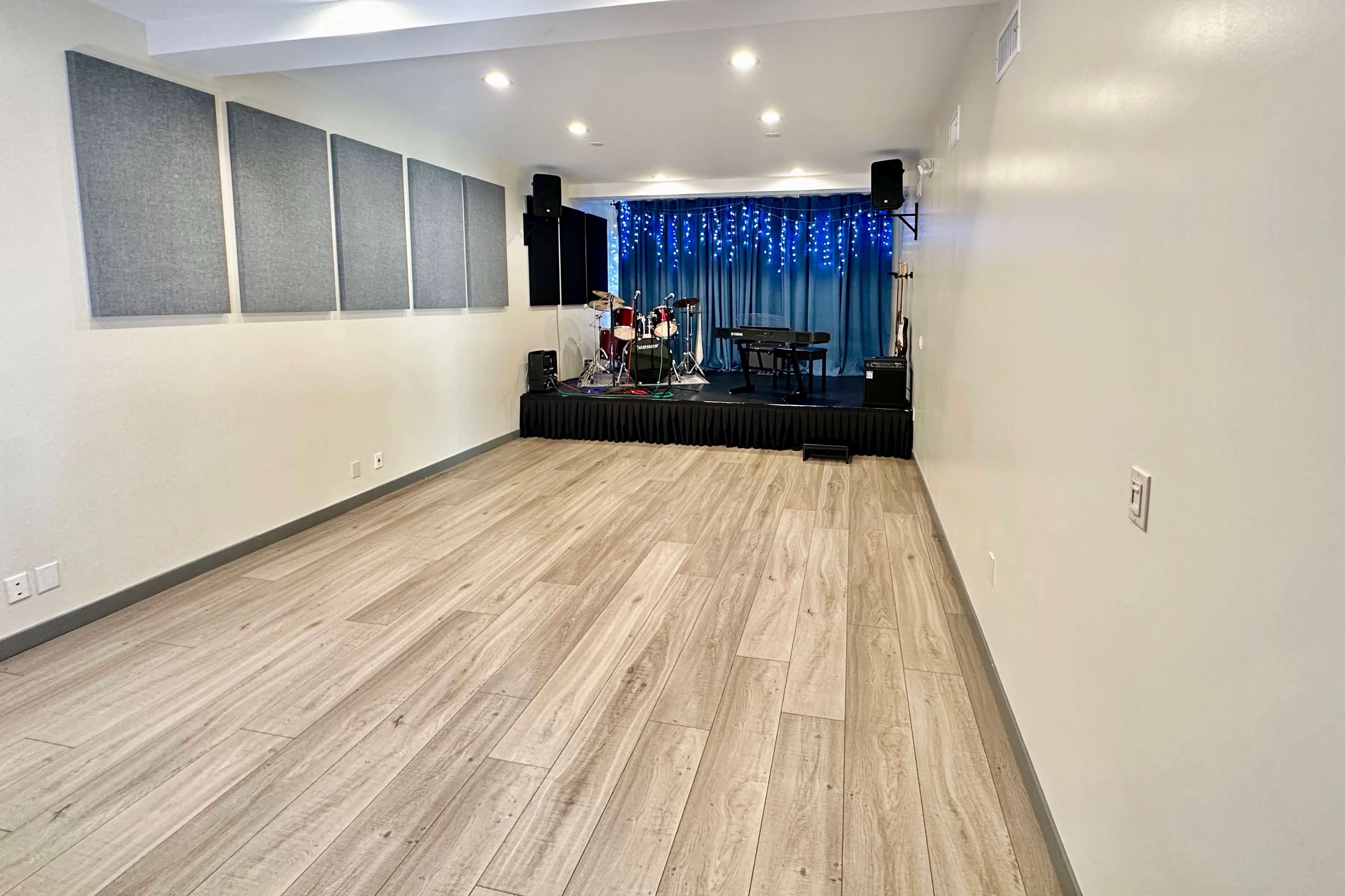 A modern room with wooden flooring, featuring a stage with musical instruments and acoustic panels on the walls.