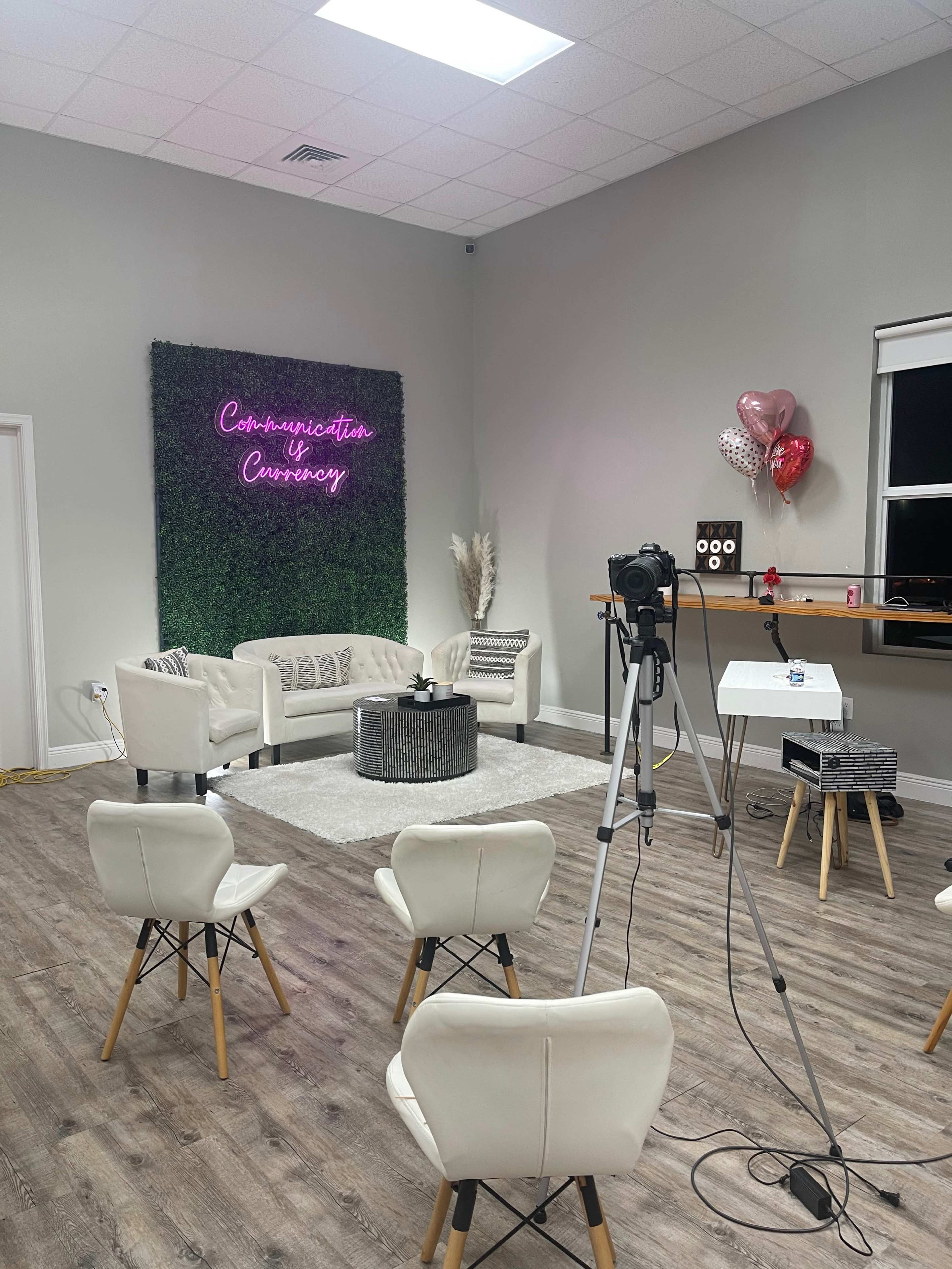 A cozy studio setup with a seating area, a neon sign on a green wall, and a camera pointed towards the seating arrangement.
