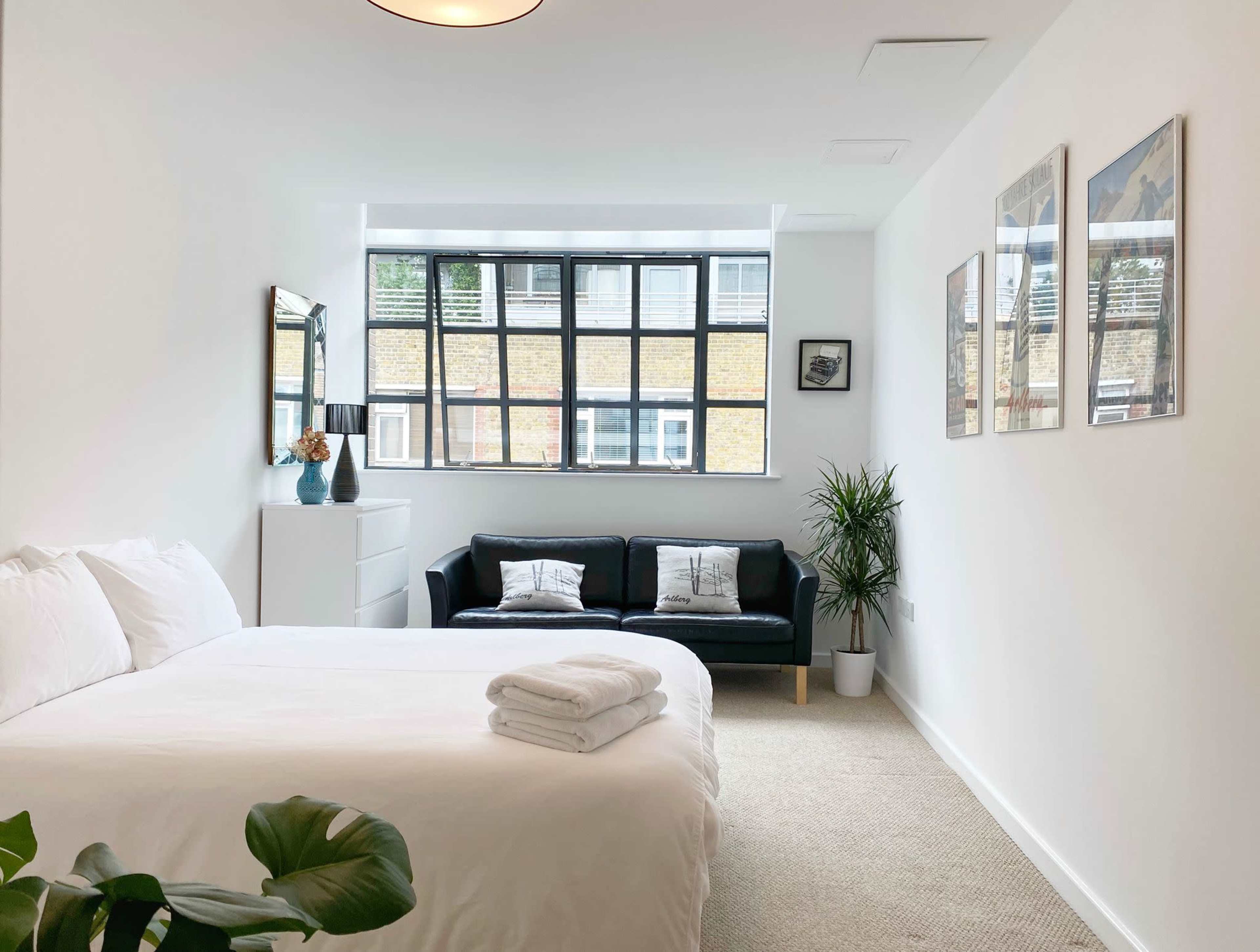 A bright bedroom with a bed, a black sofa, a small plant, and framed pictures on the white walls.
