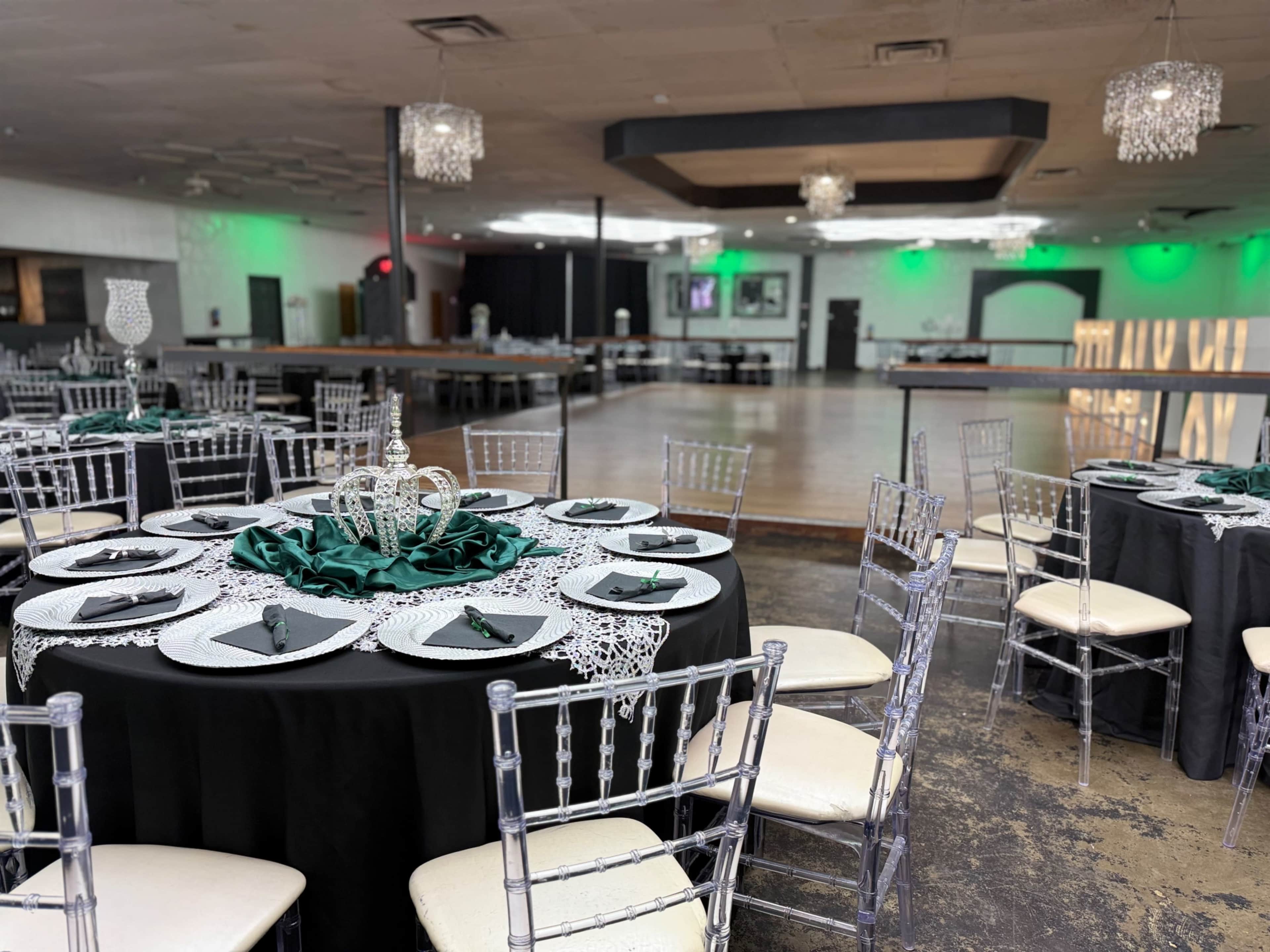 A formal dining area features a round table set with dark green and silver decorations, surrounded by clear chiavari chairs, in a spacious venue with dim lighting and a polished dance floor.