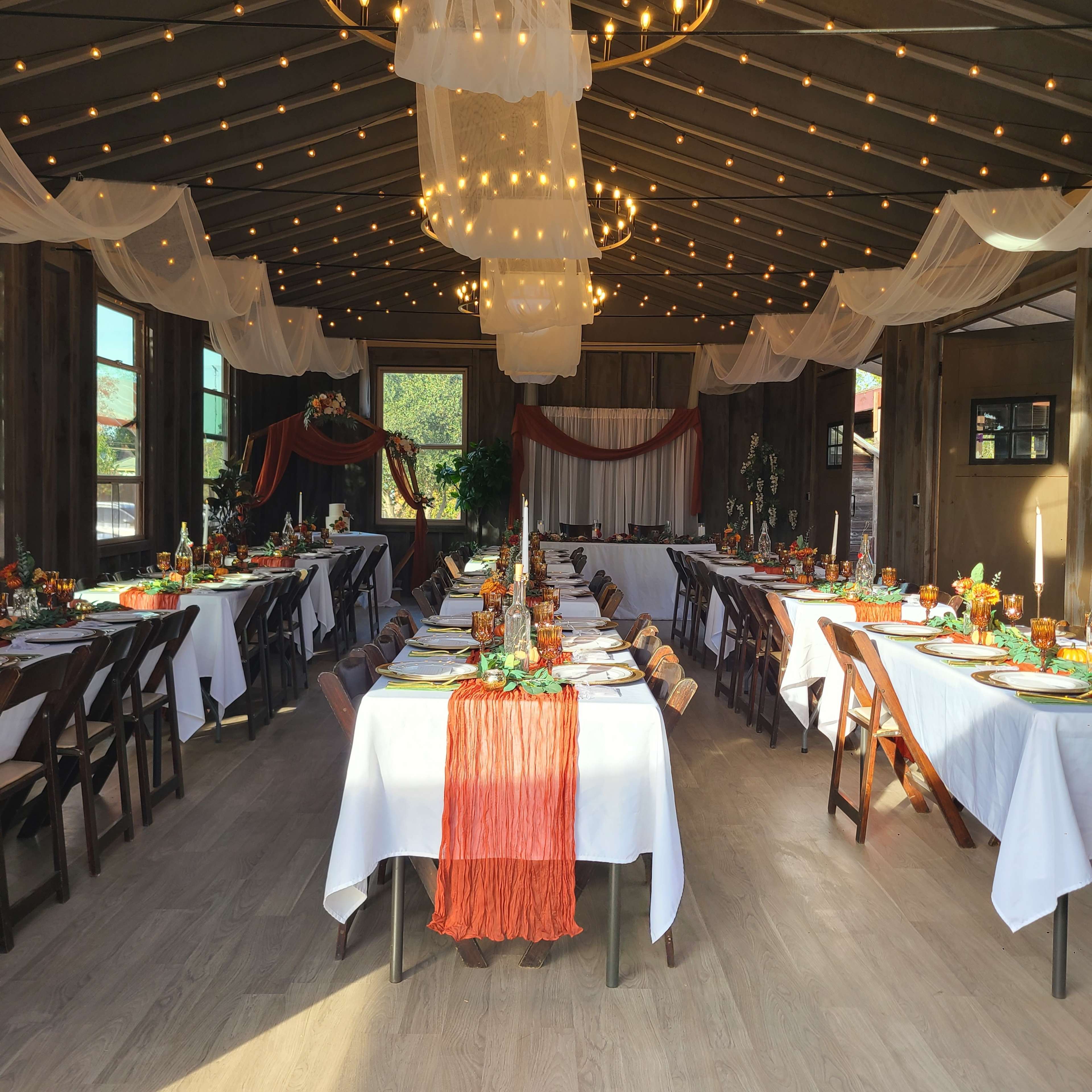 The image shows a beautifully arranged dining area with multiple tables set for an event, featuring white tablecloths, colorful centerpieces, and decorative lighting.
