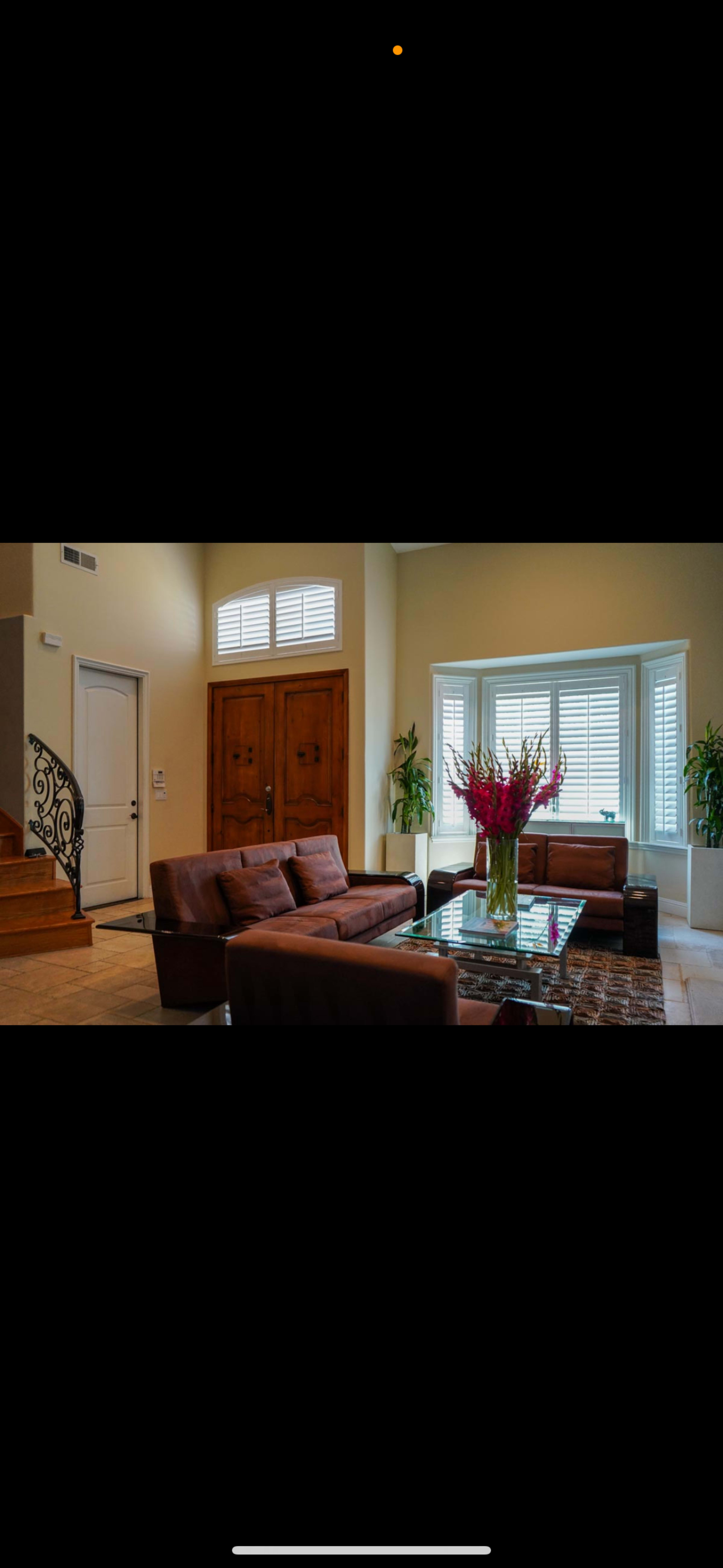 A modern living room features two brown sofas, a glass coffee table, and large windows with plants nearby, set against a backdrop of a staircase and decorative doors.