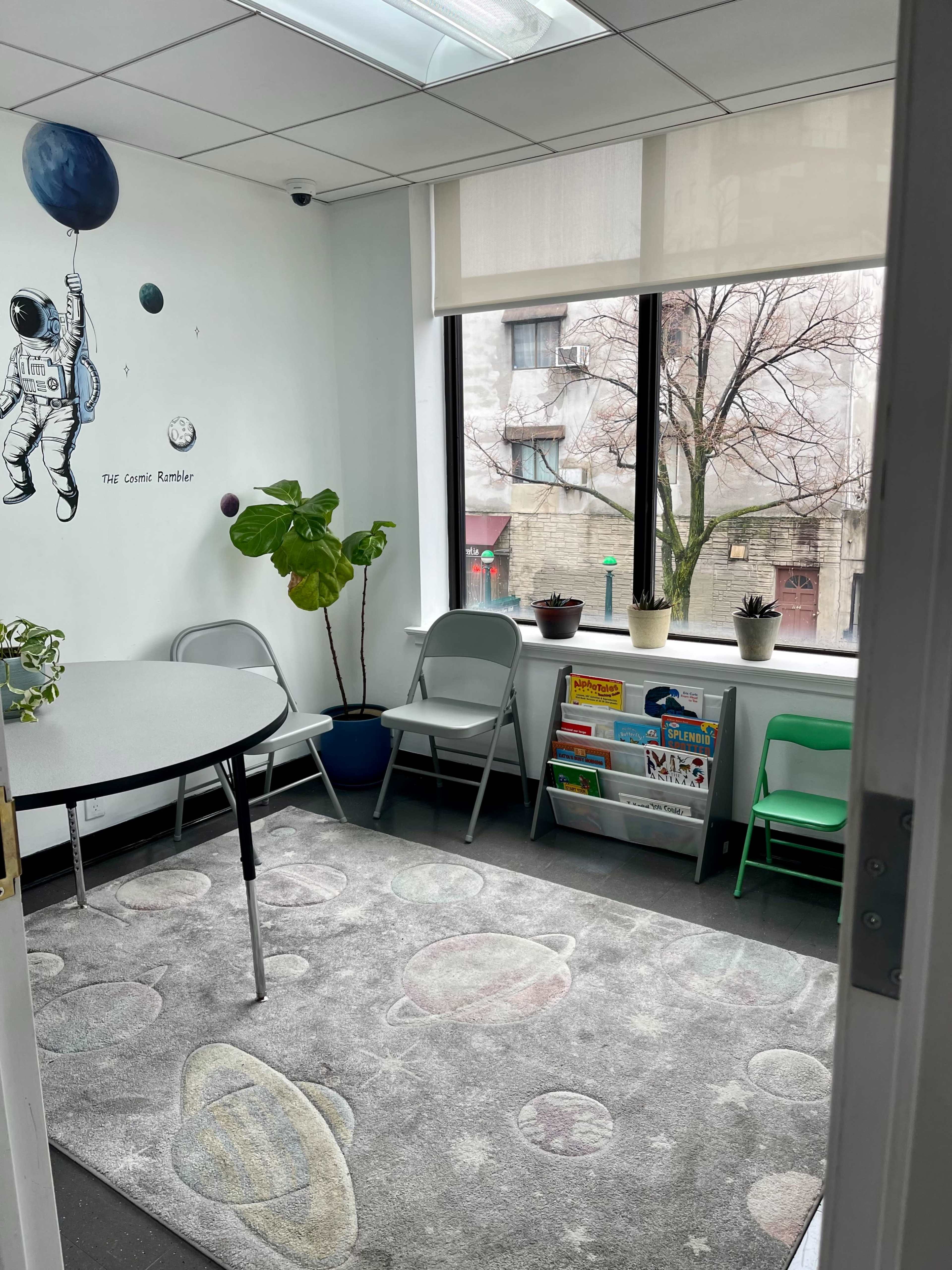 A small, brightly-lit room features a round table, chairs, a rug with a space theme, and a window overlooking a tree-lined street.