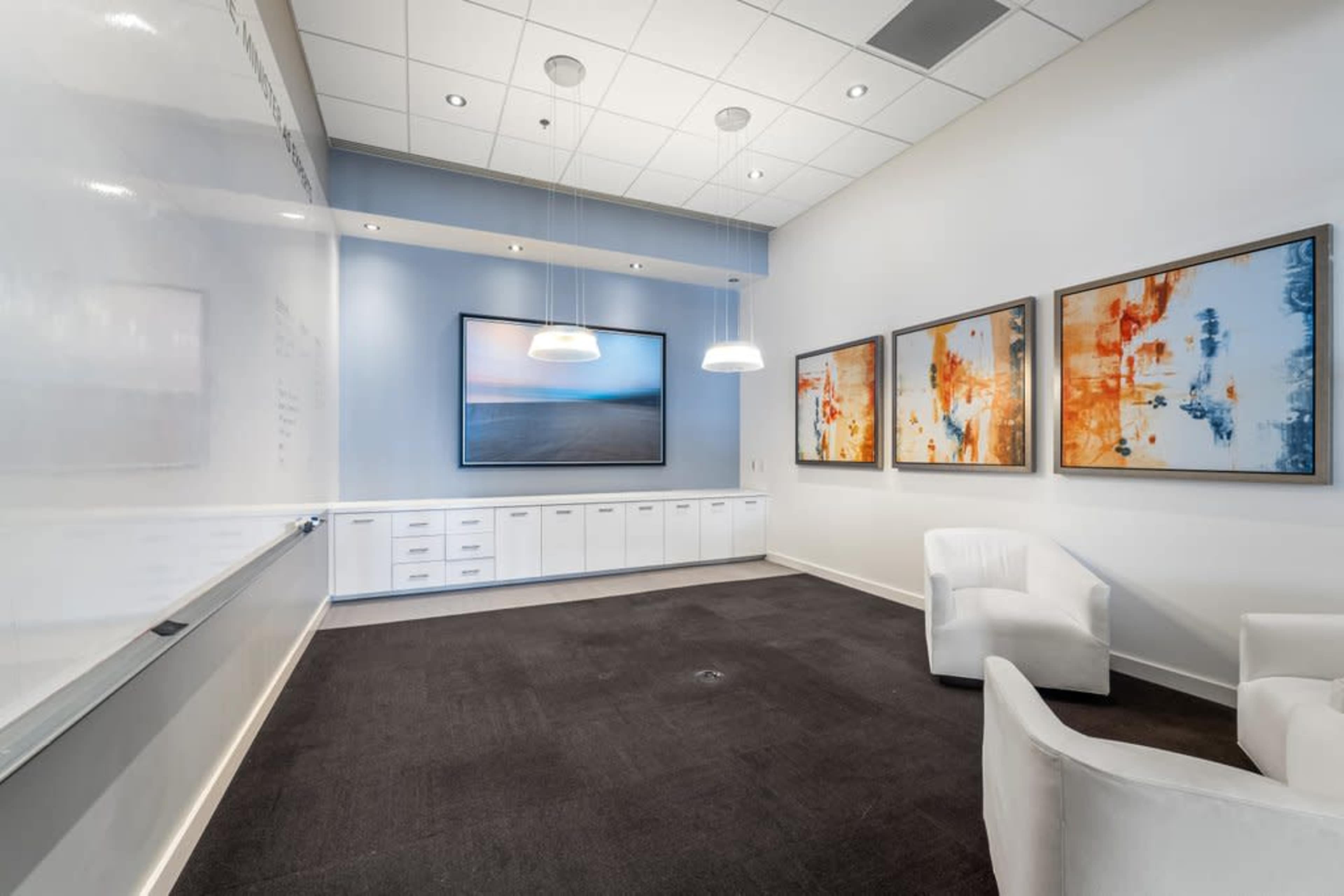 The image shows a modern meeting room featuring a large whiteboard, a digital screen displaying an image, and three abstract paintings on the walls, with two white chairs arranged in the space.