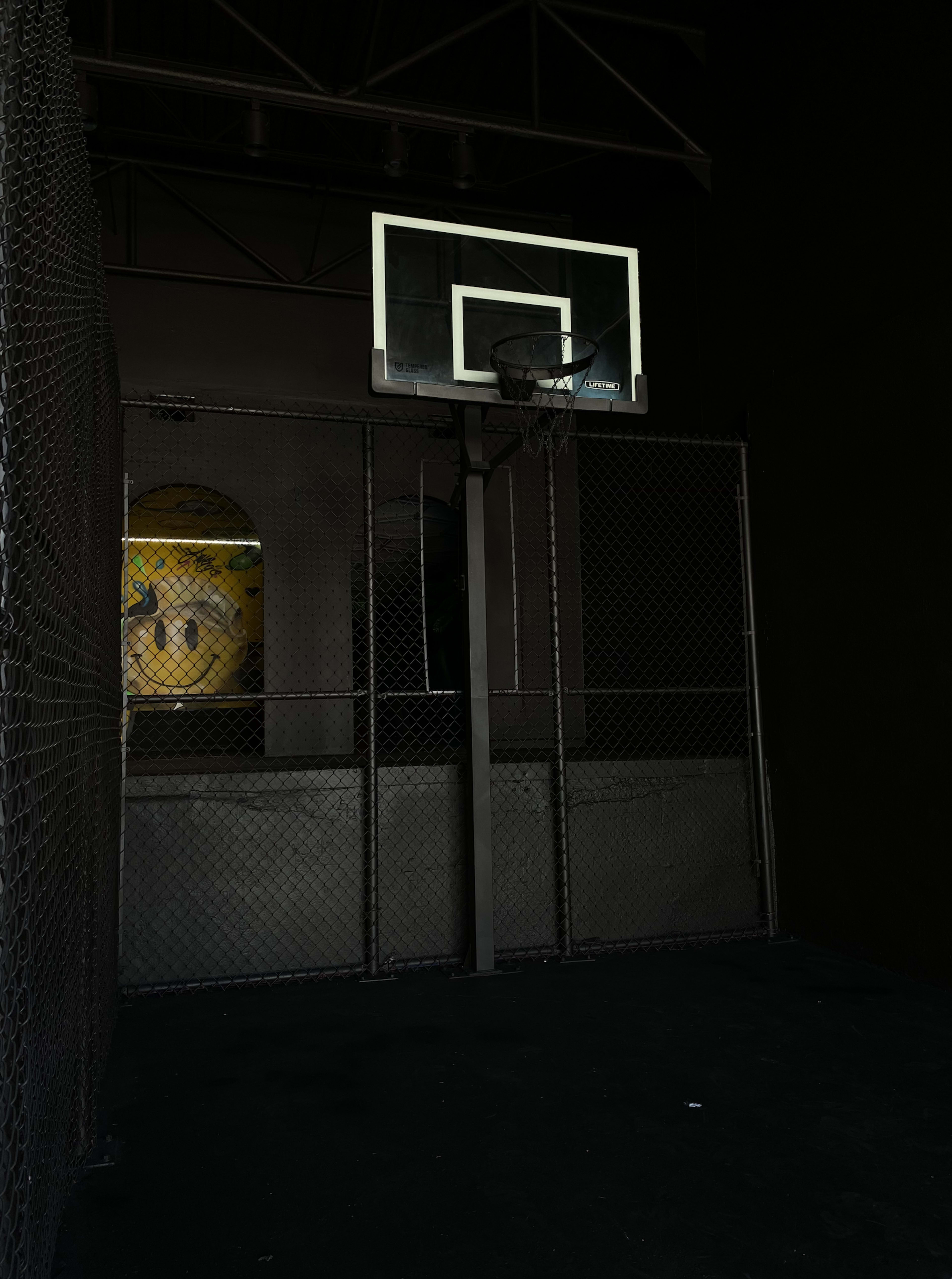 A basketball hoop attached to a black wall within a fenced area, with a large, colorful smiley face mural visible in the background.