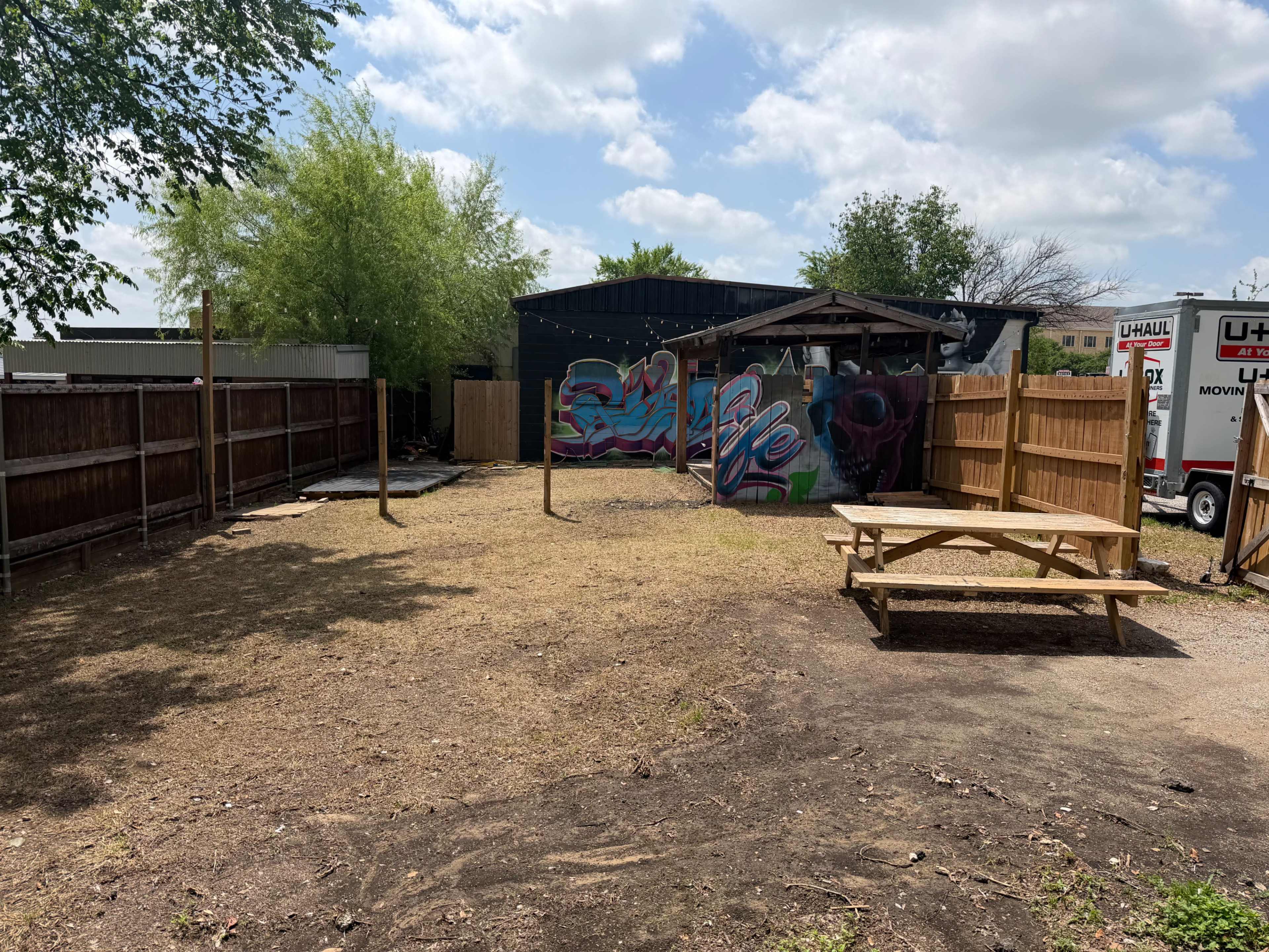 The scene depicts an outdoor space with a picnic table, wooden fencing, a mural on a building, and several wooden posts.