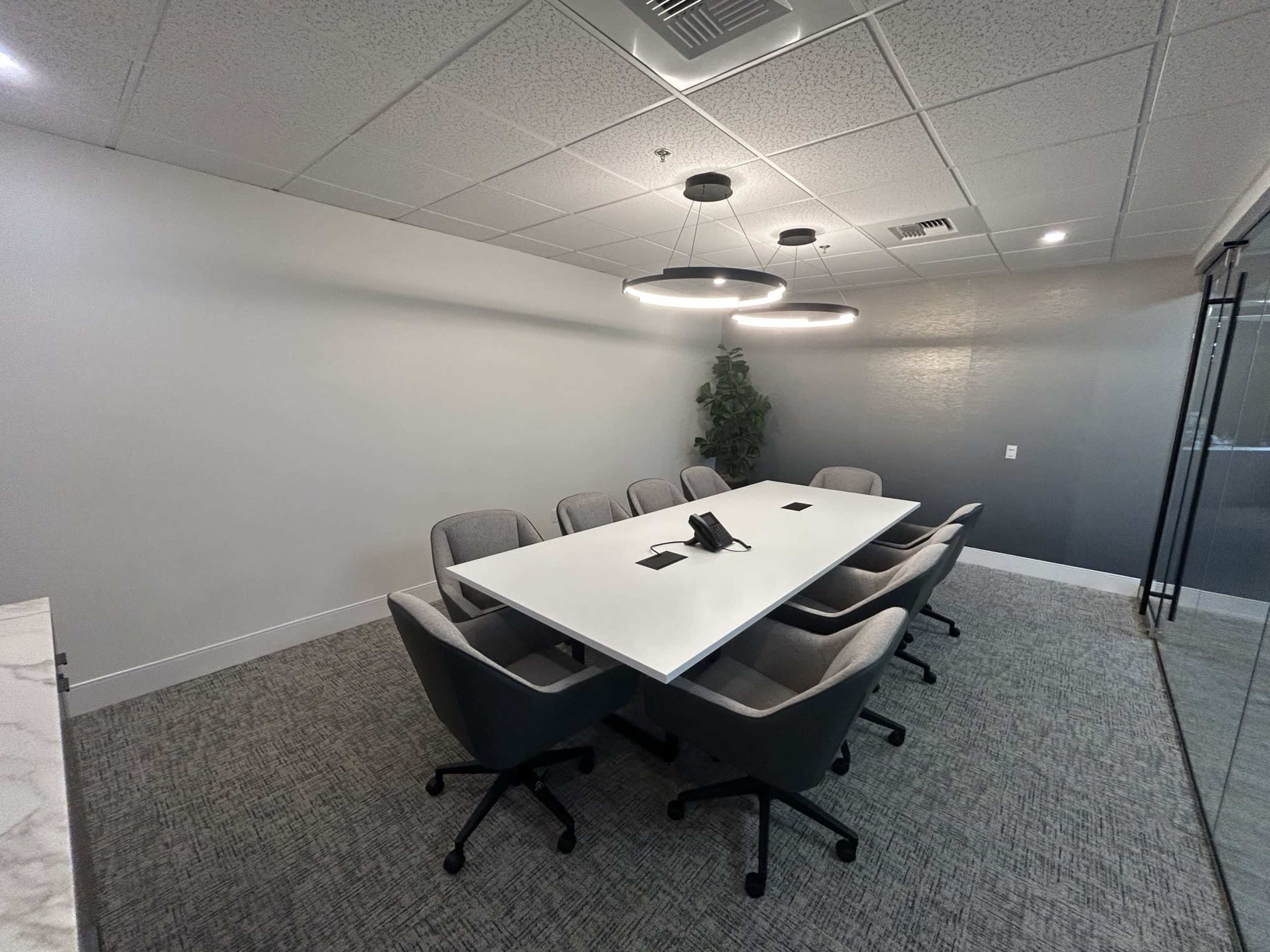 A modern conference room features a large white table surrounded by ergonomic chairs, with a plant in the corner and two circular overhead lights.