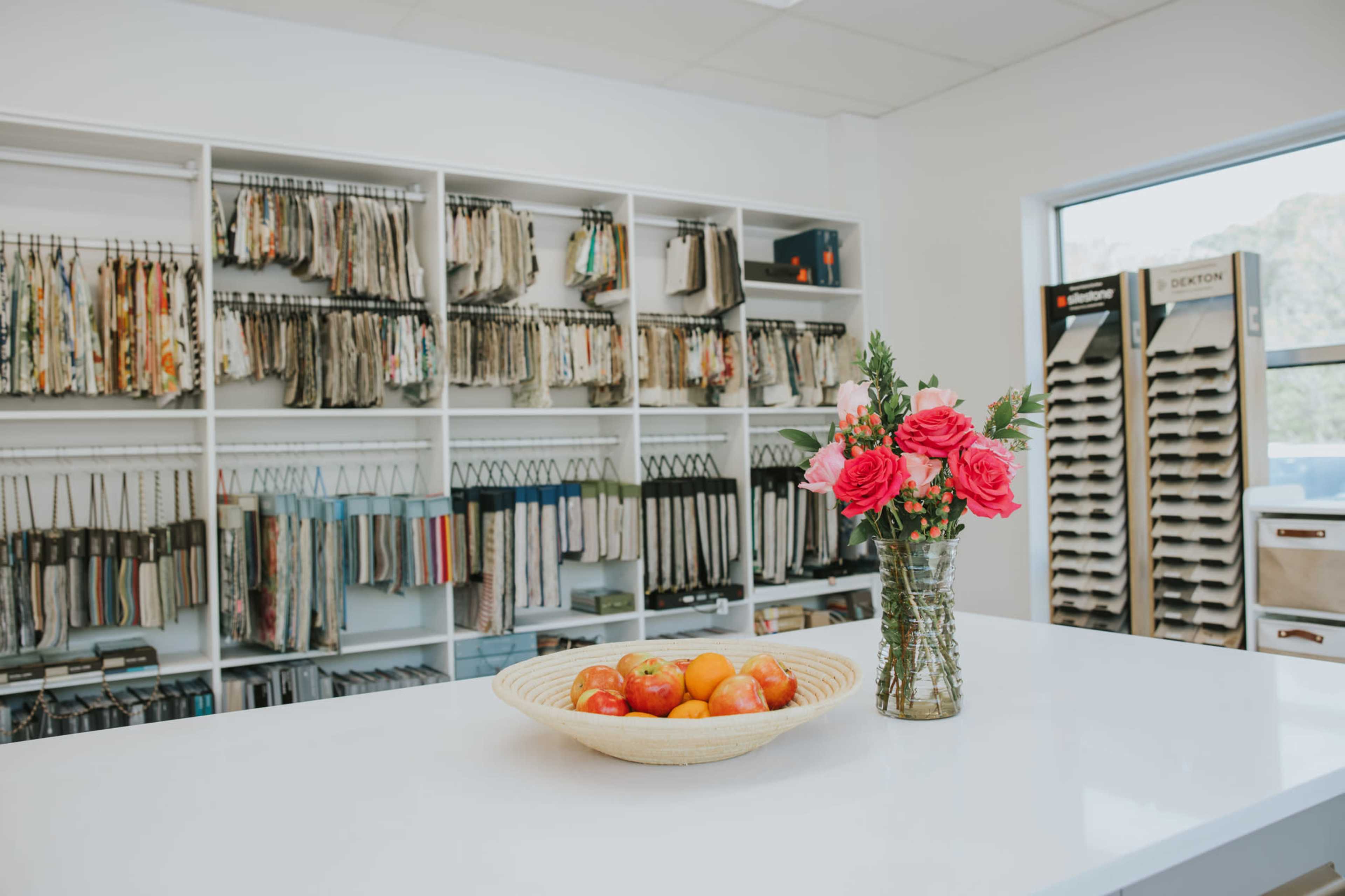 A white countertop displays a basket of fruit and a vase of roses, while fabric samples hang on shelves in the background.