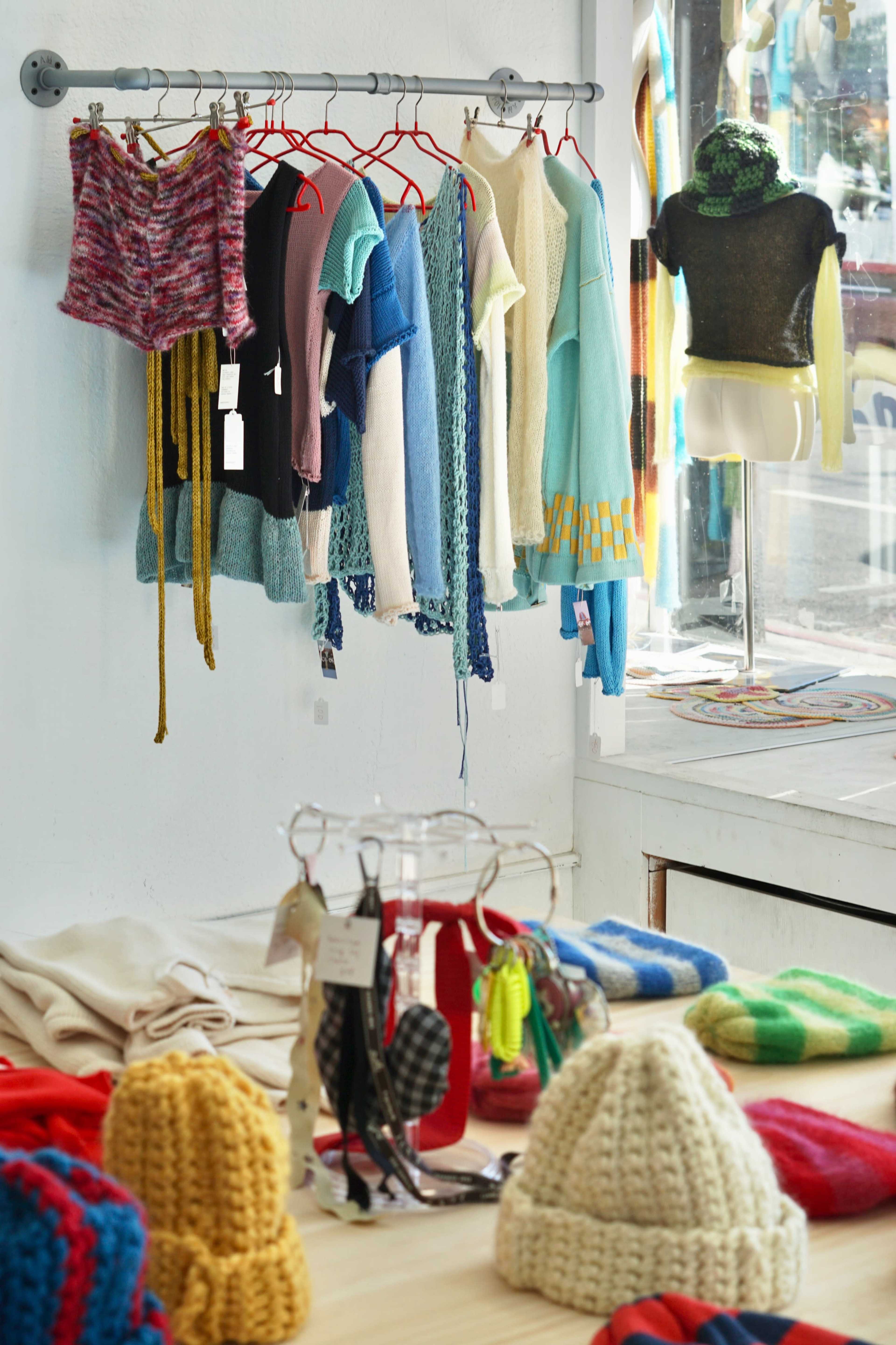 A clothing rack displays a variety of colorful knitted garments, while a table in the foreground holds several knitted hats and accessories.