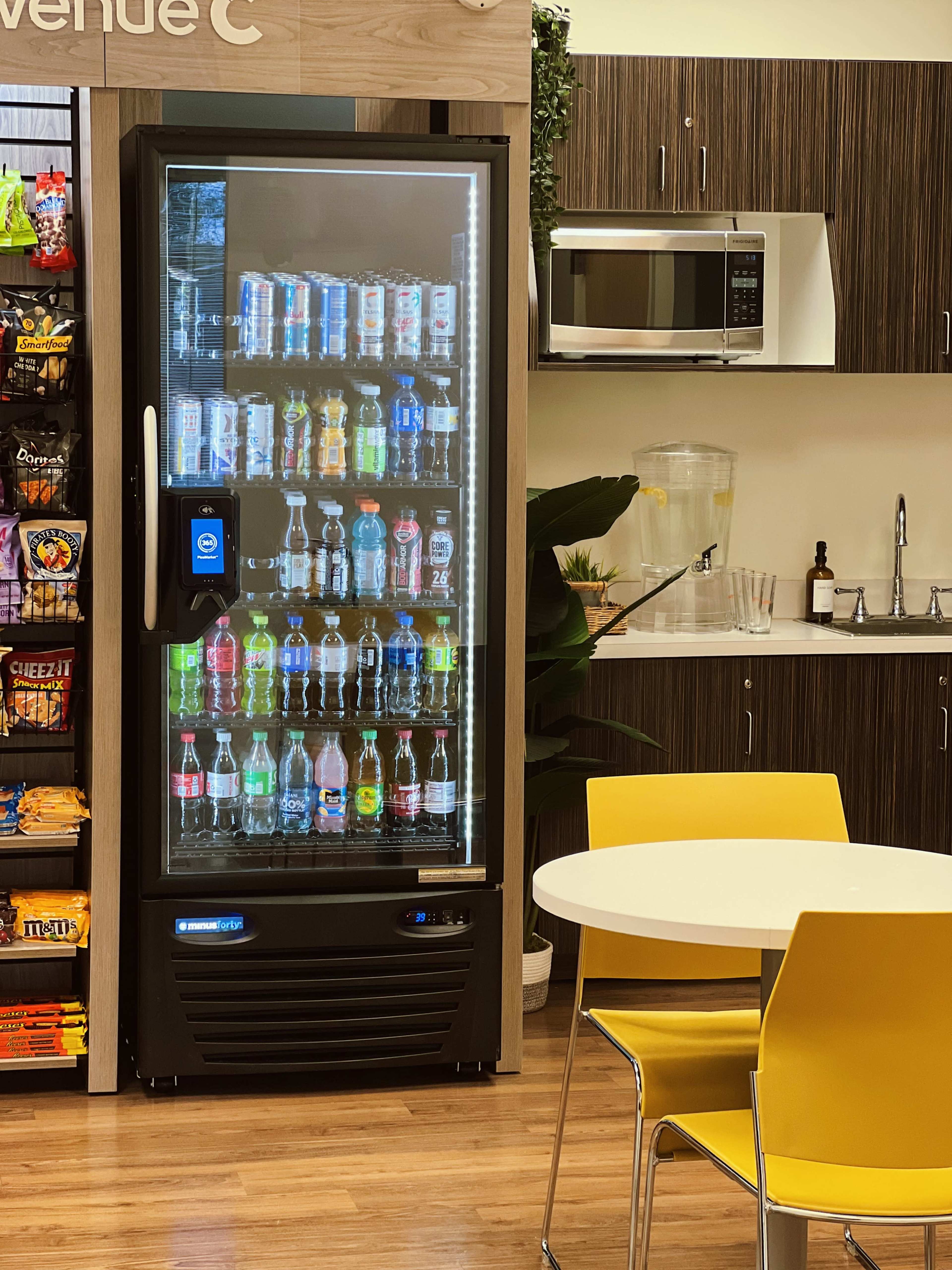 A vending machine filled with various bottled beverages is positioned next to a kitchenette featuring a microwave and a table with yellow chairs.