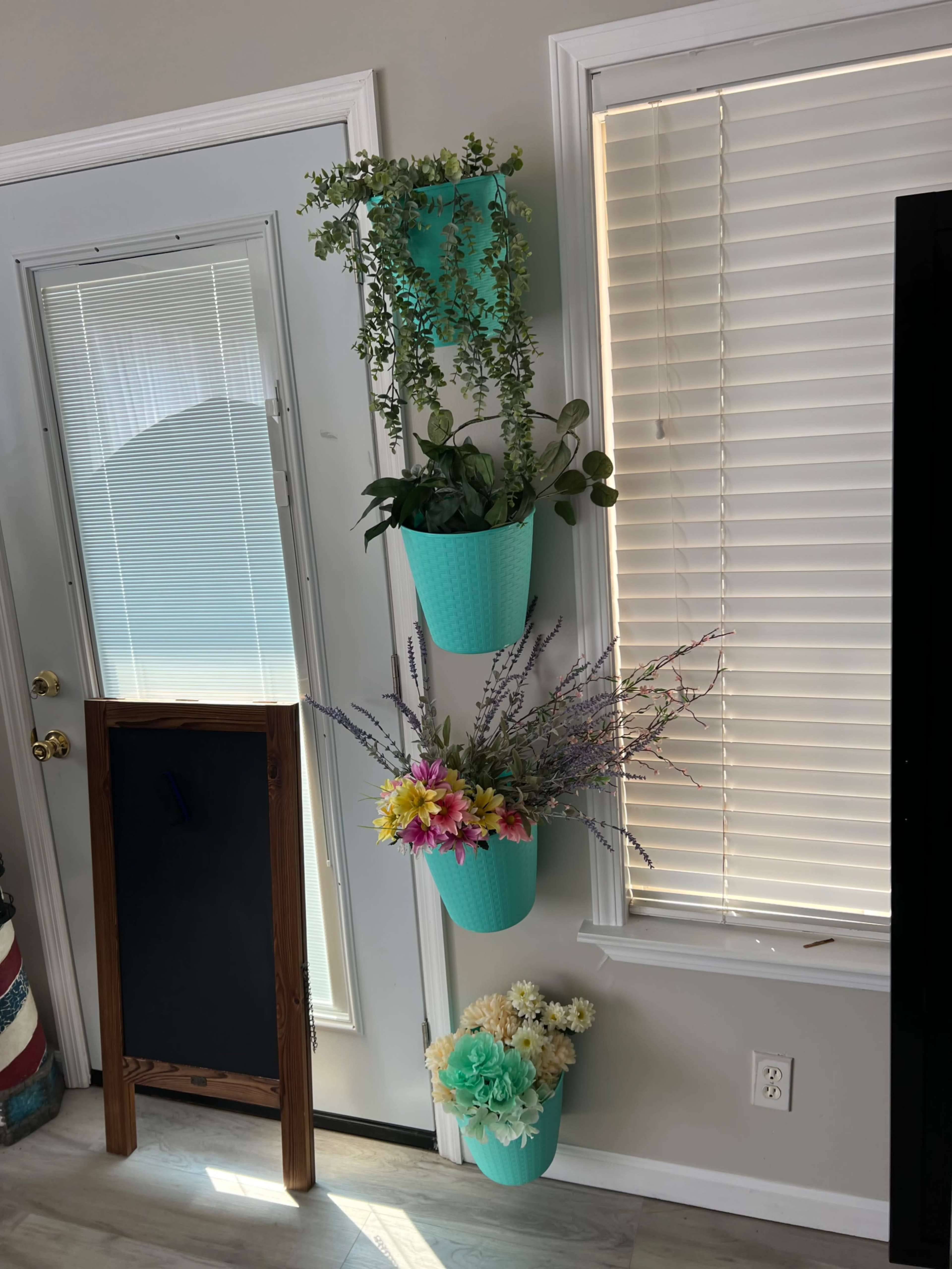 Three turquoise planters, stacked vertically on a wall, contain various plants and artificial flowers near a door with blinds.