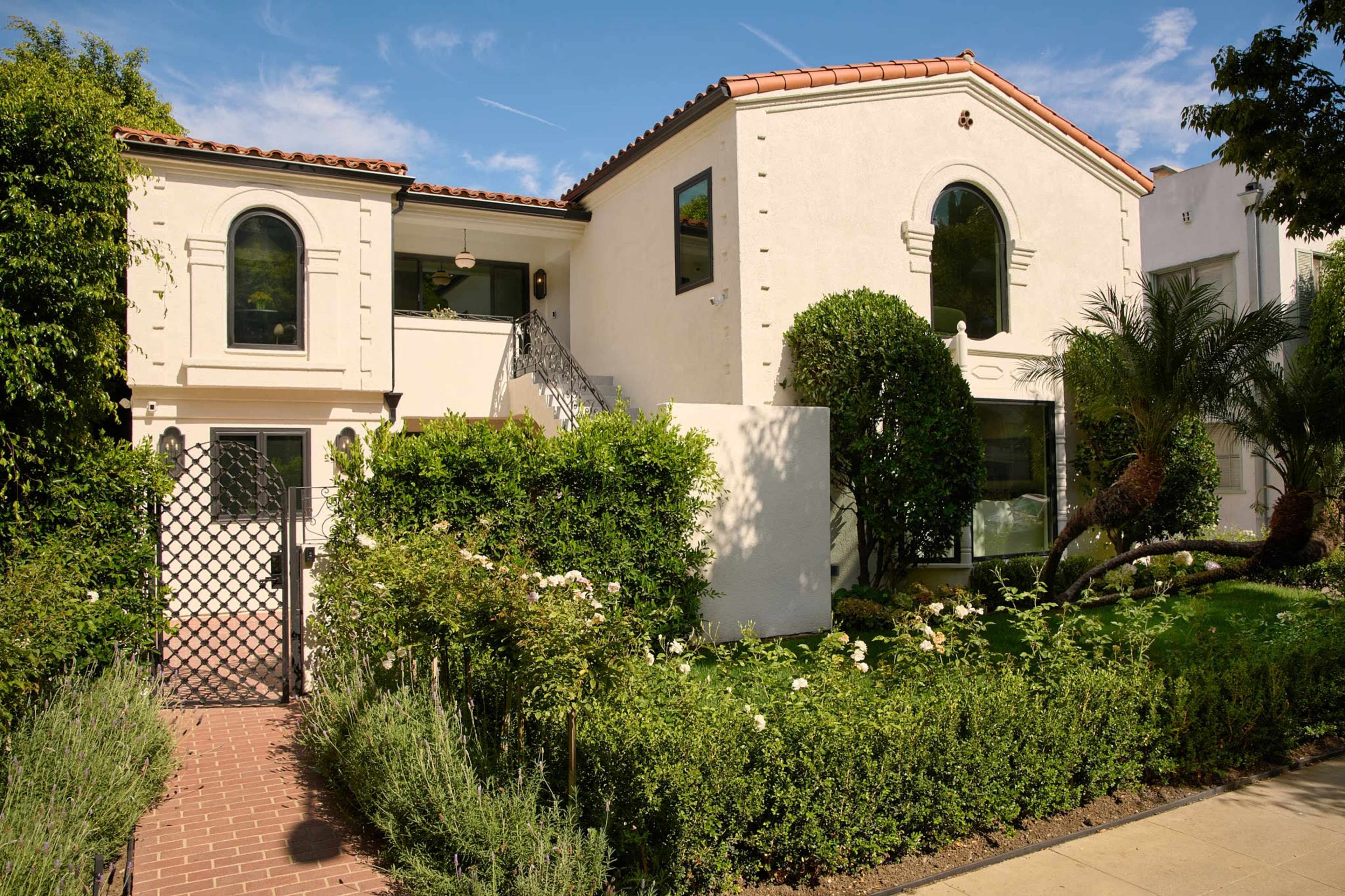 The image shows a two-story, white stucco house with arched windows and a tiled roof, surrounded by greenery and a well-maintained garden.