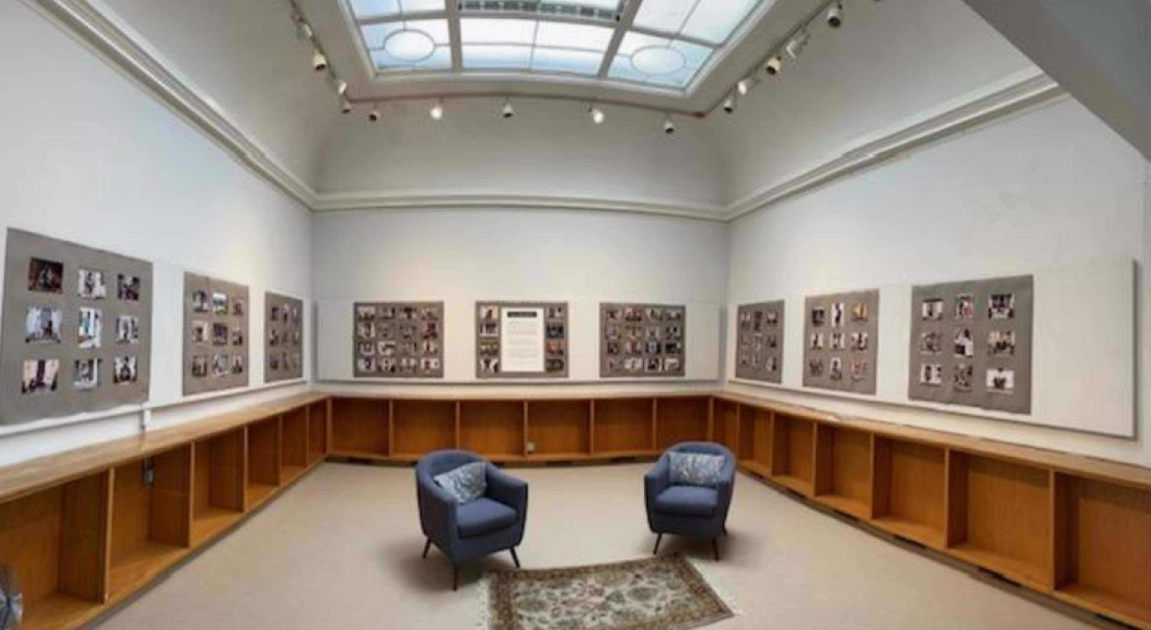 A gallery room with two blue chairs situated on a carpet, surrounded by walls displaying framed photographs.