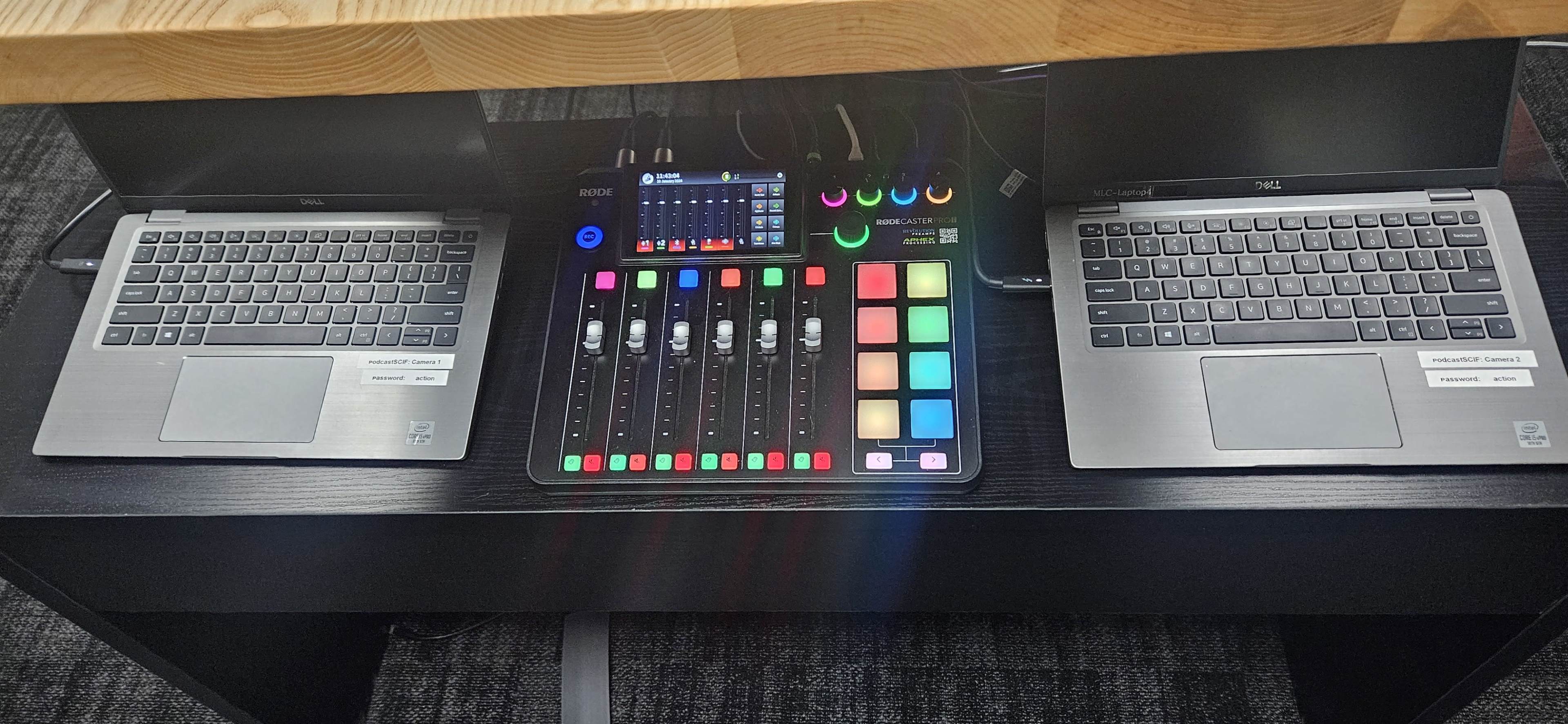 The image shows a workspace with two laptops on either side of a control surface featuring faders and colorful buttons.
