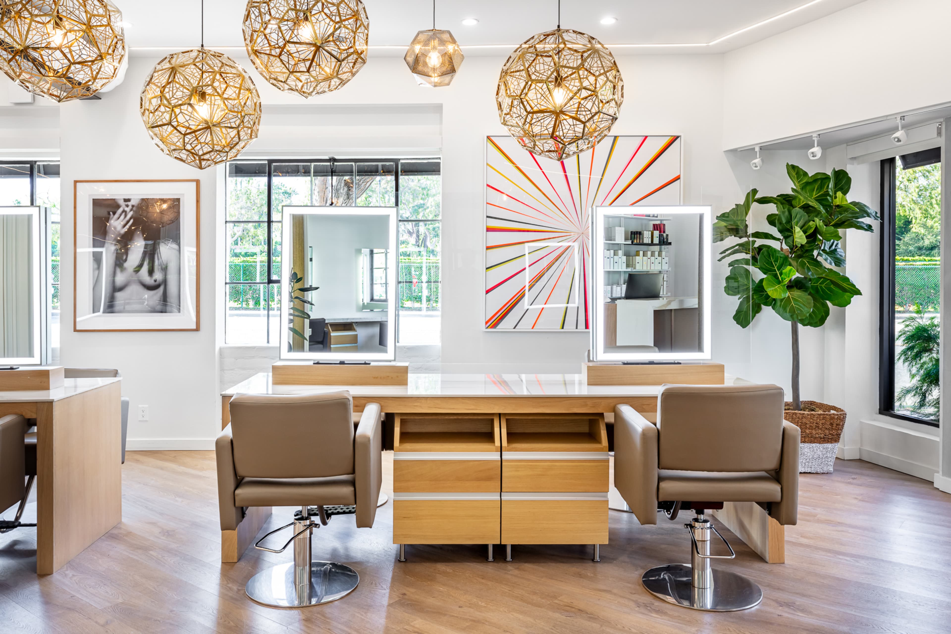 A modern salon interior featuring two styling stations with mirrors, wooden furniture, decorative hanging lights, and large windows with views of greenery.