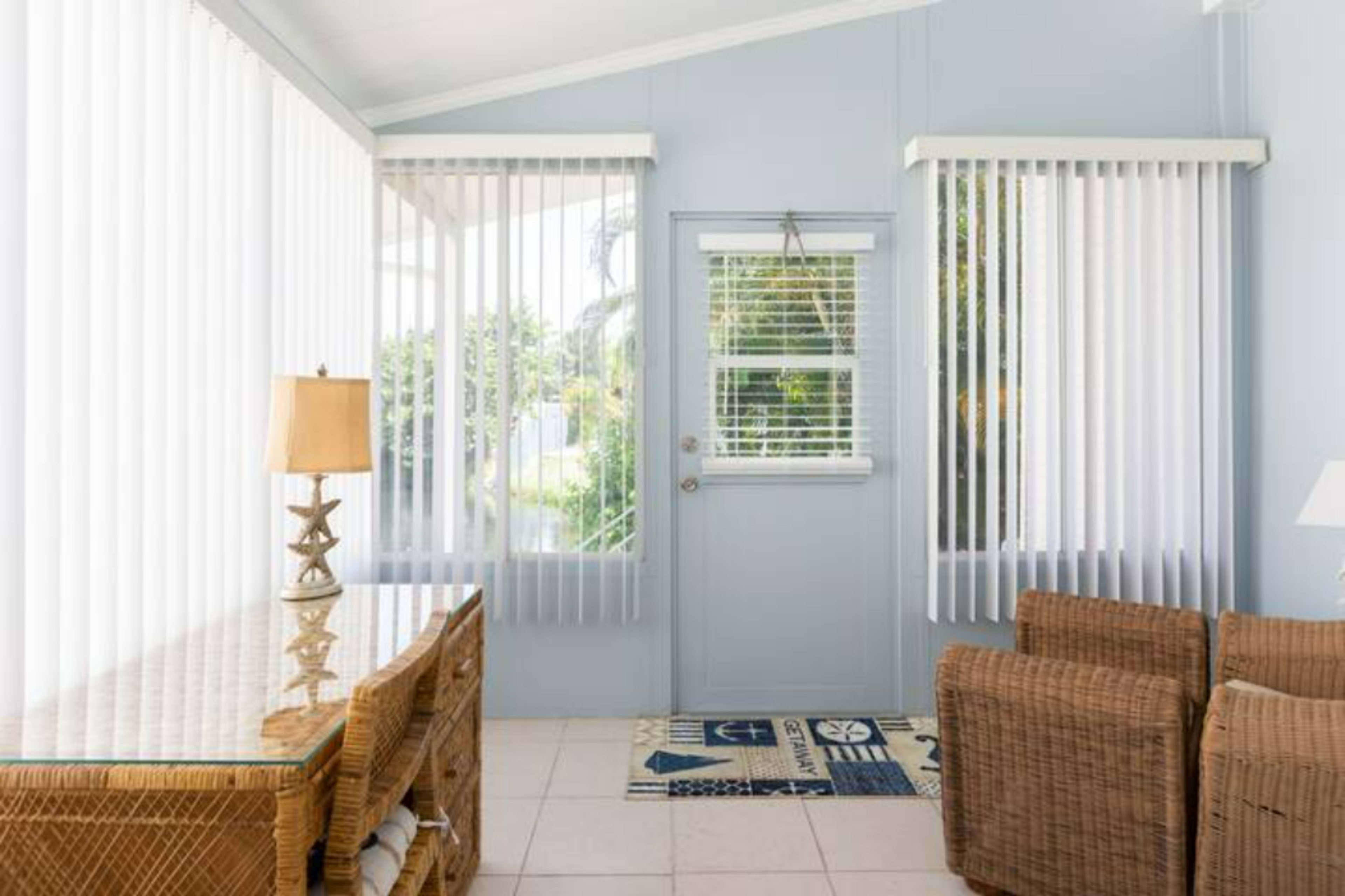 A sunlit room features a glass door and large windows adorned with vertical blinds, alongside a wicker coffee table and two chairs.