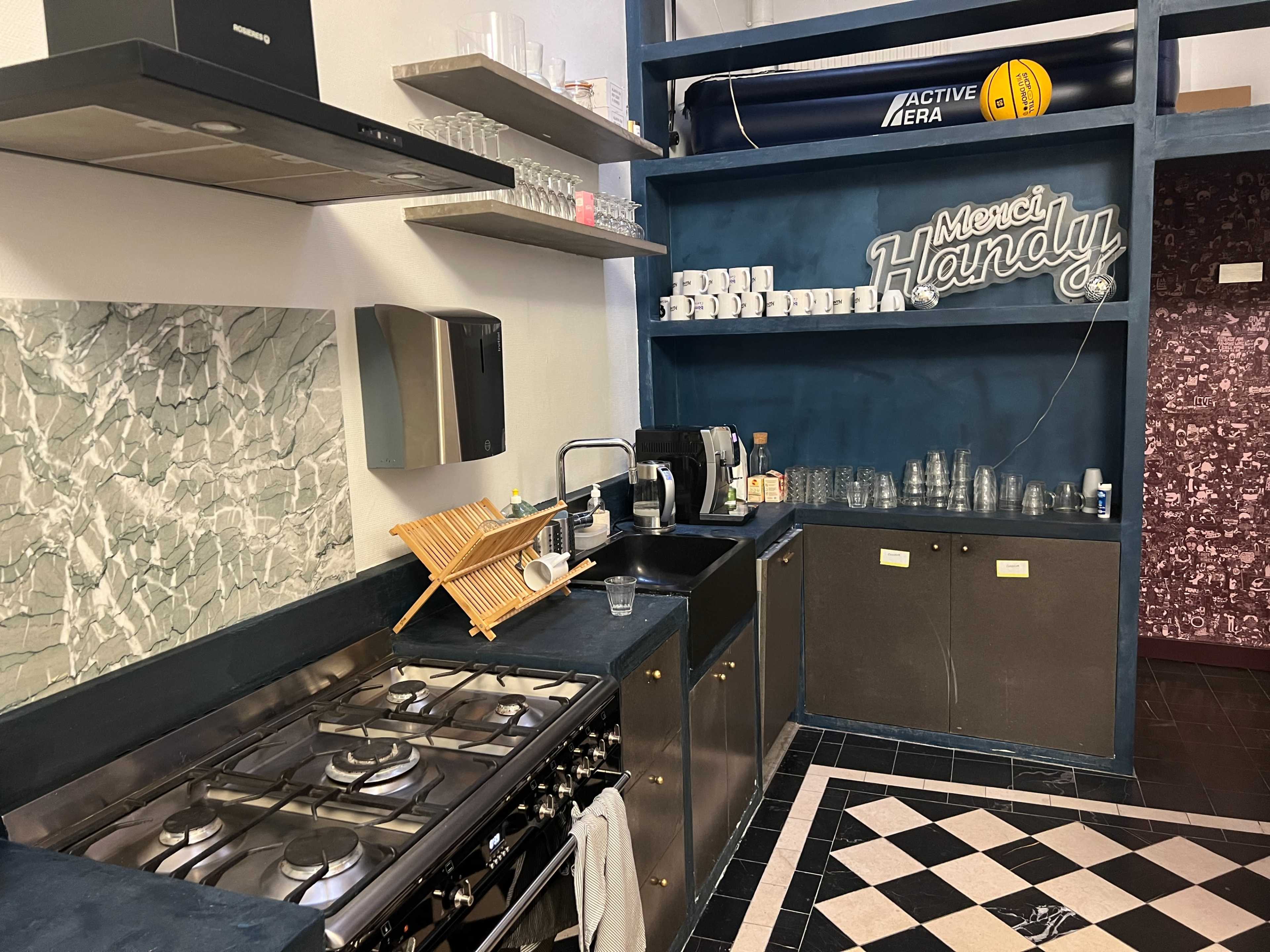 The image shows a modern kitchen with a gas stove, sink, and shelves displaying various glassware and utensils.