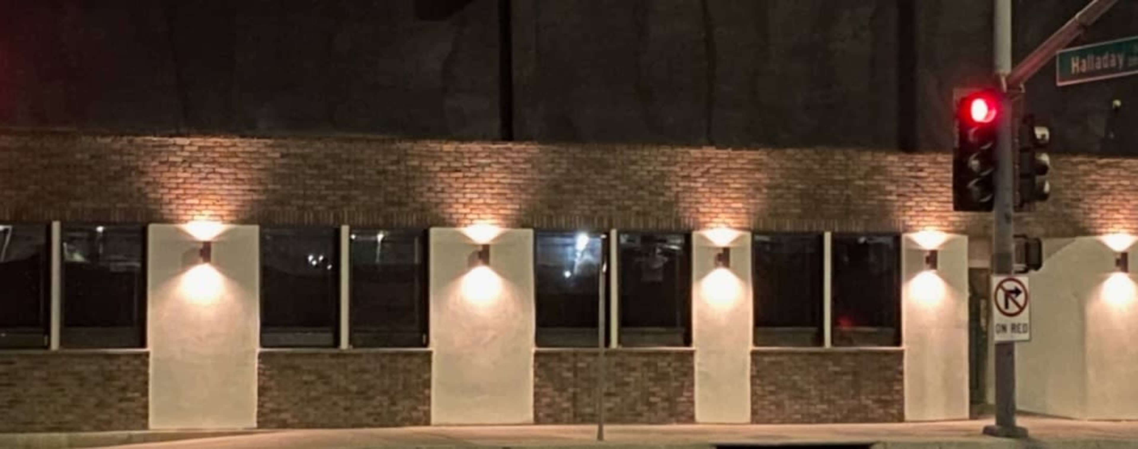 A brick building with illuminated wall lights and large windows stands at an intersection with a red traffic signal.