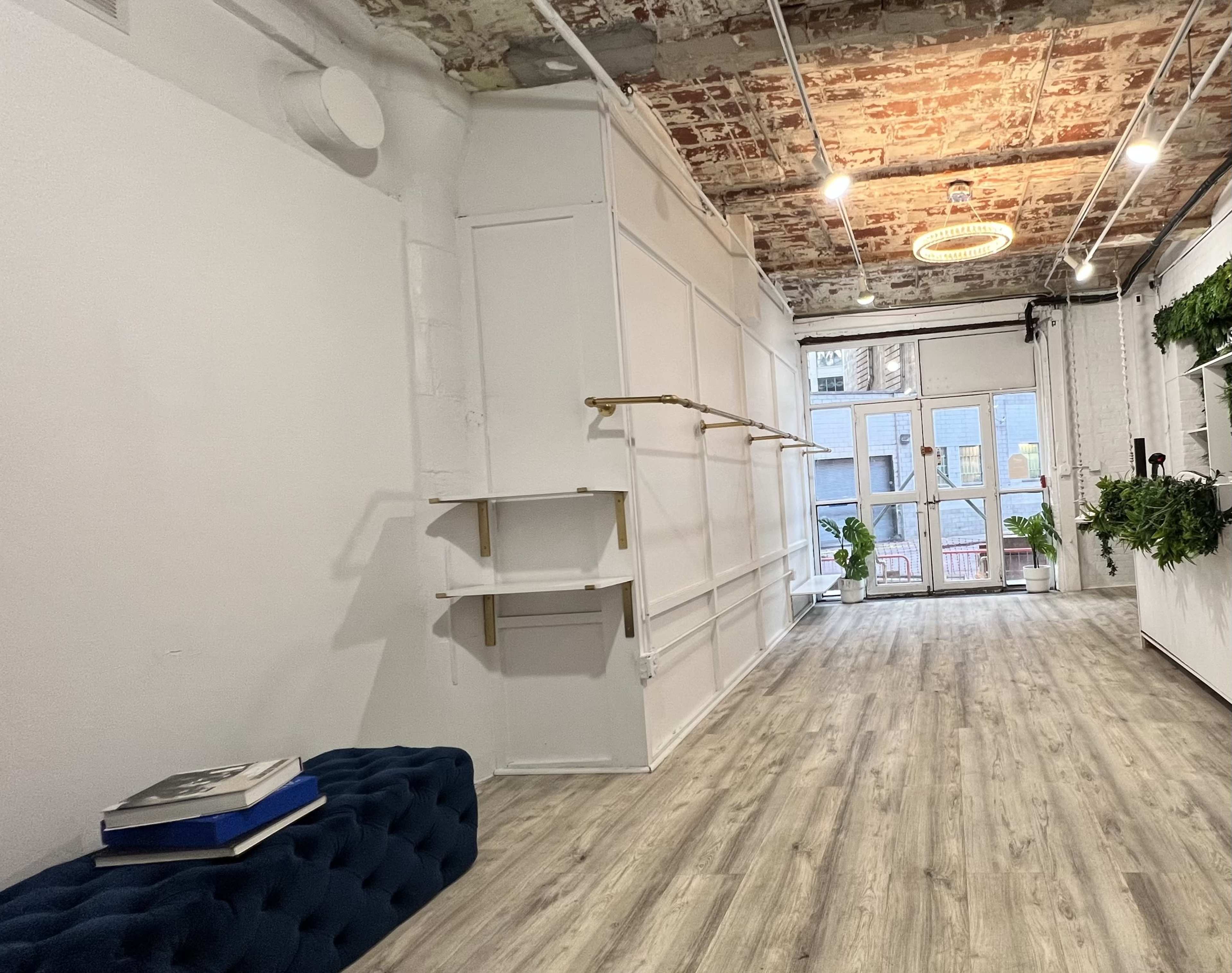 The image shows a spacious interior with wooden flooring, a brick ceiling, and minimalist decor, featuring a blue upholstered bench and potted plants near glass doors.
