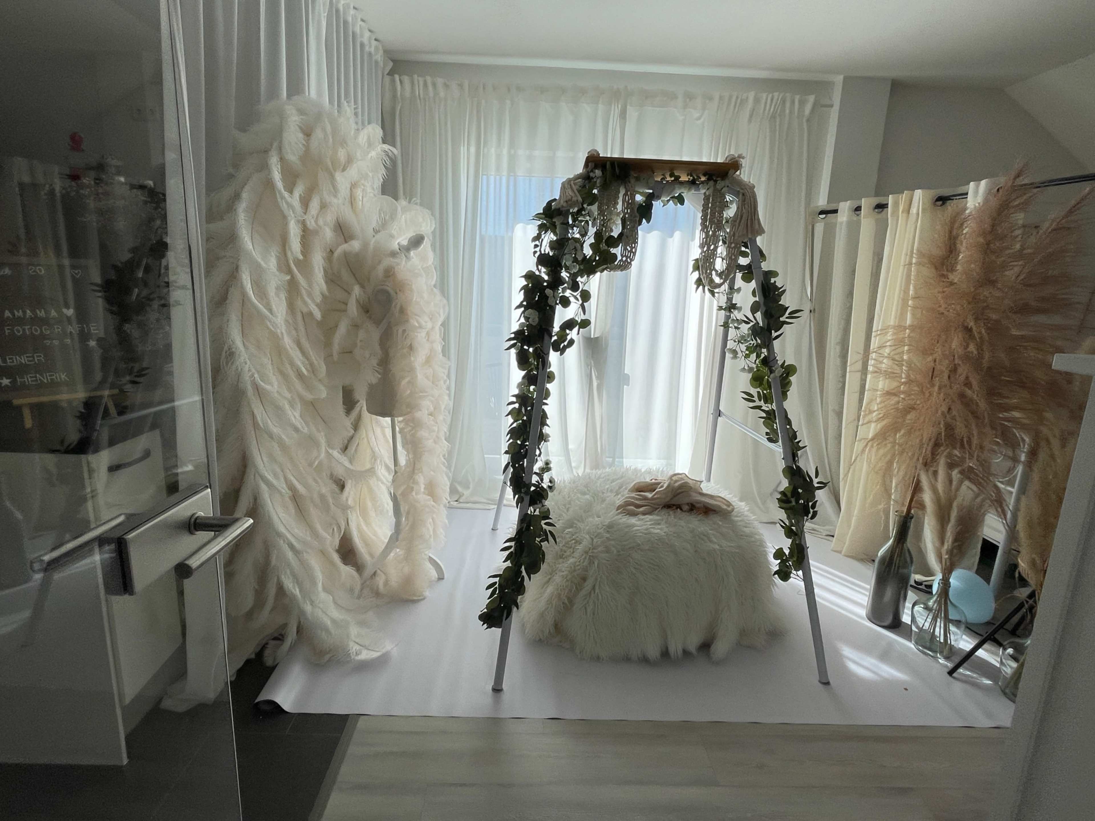 The image shows a well-lit room with a plush, white bed surrounded by decorative greenery and soft fabric drapes.