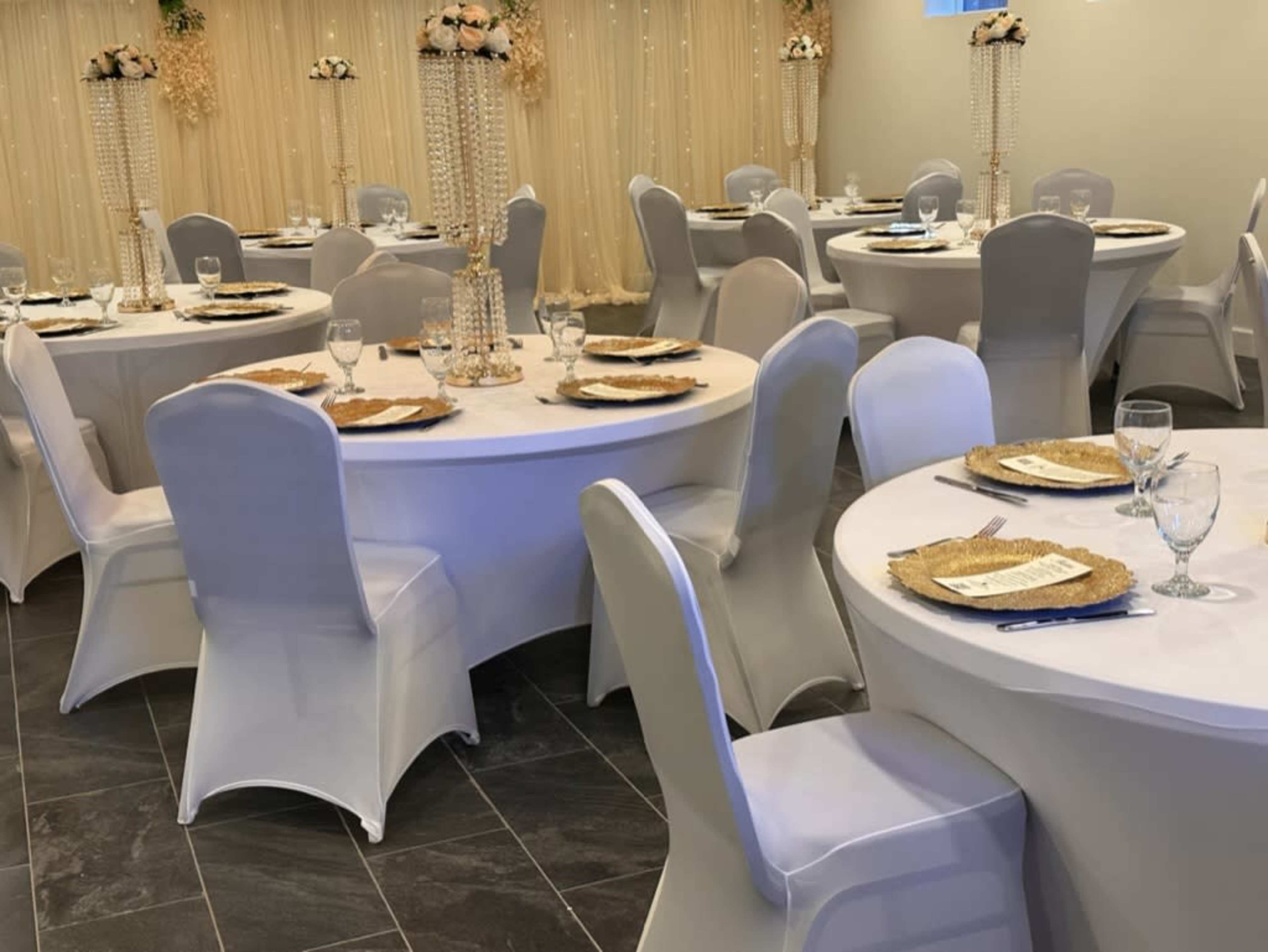 The image shows a banquet hall set up with round tables covered in white tablecloths, each arranged with plates and glassware, surrounded by elegant décor.