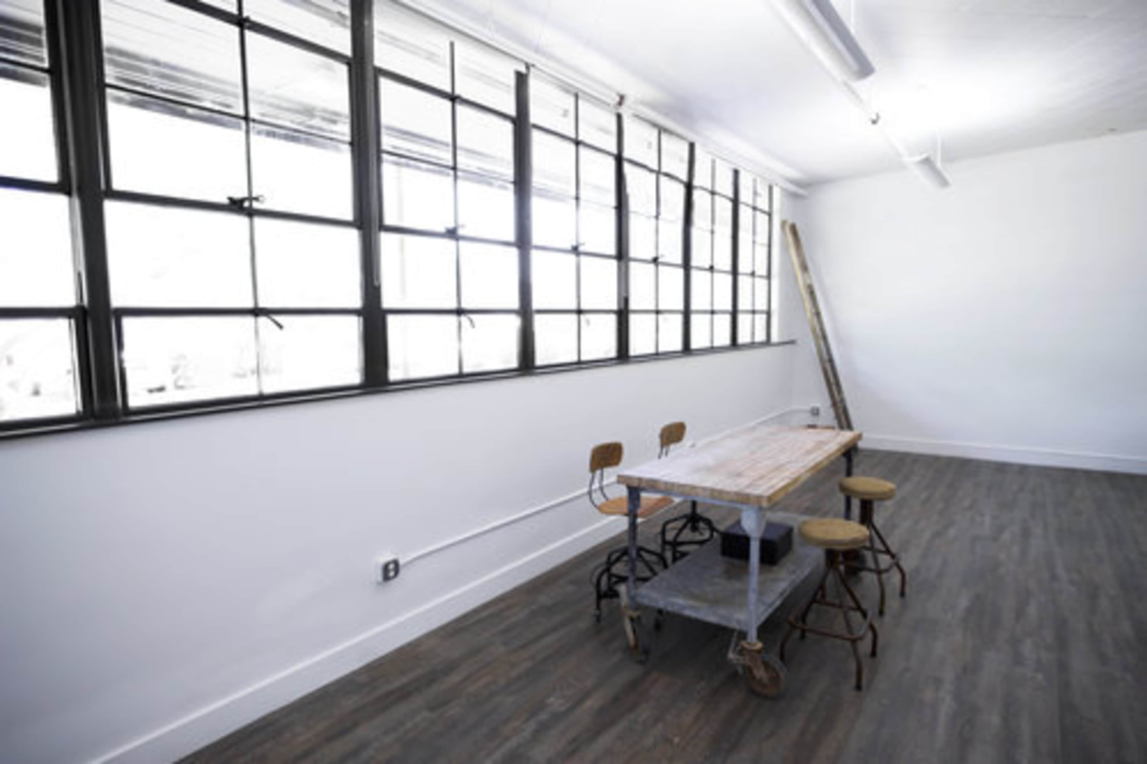 A minimalist room with large windows, a wooden table on wheels, and four metal chairs arranged around it.