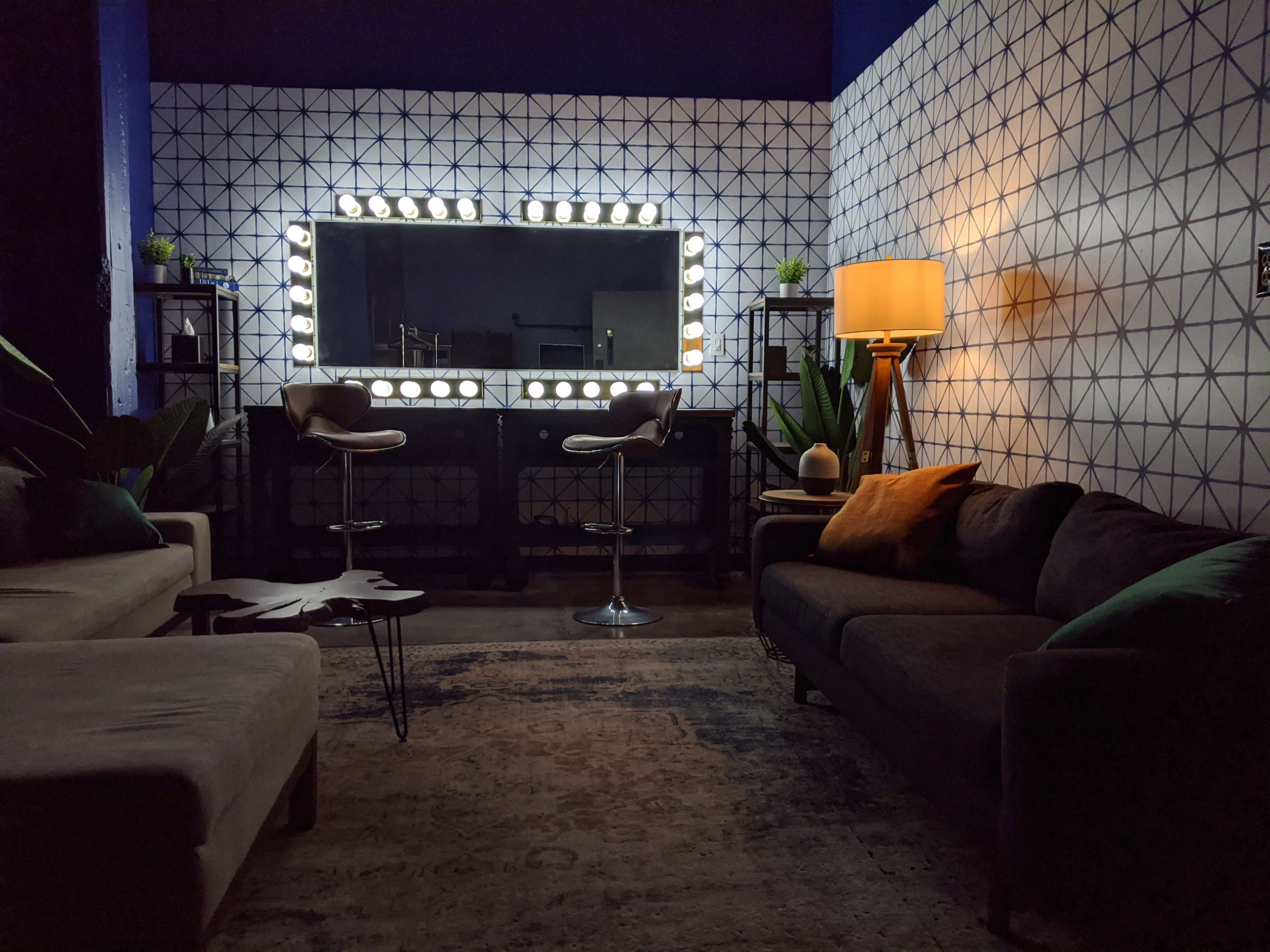 The image shows a stylish lounge area featuring a large illuminated mirror, two bar stools, a floor lamp, and two sofas arranged around a decorative table.