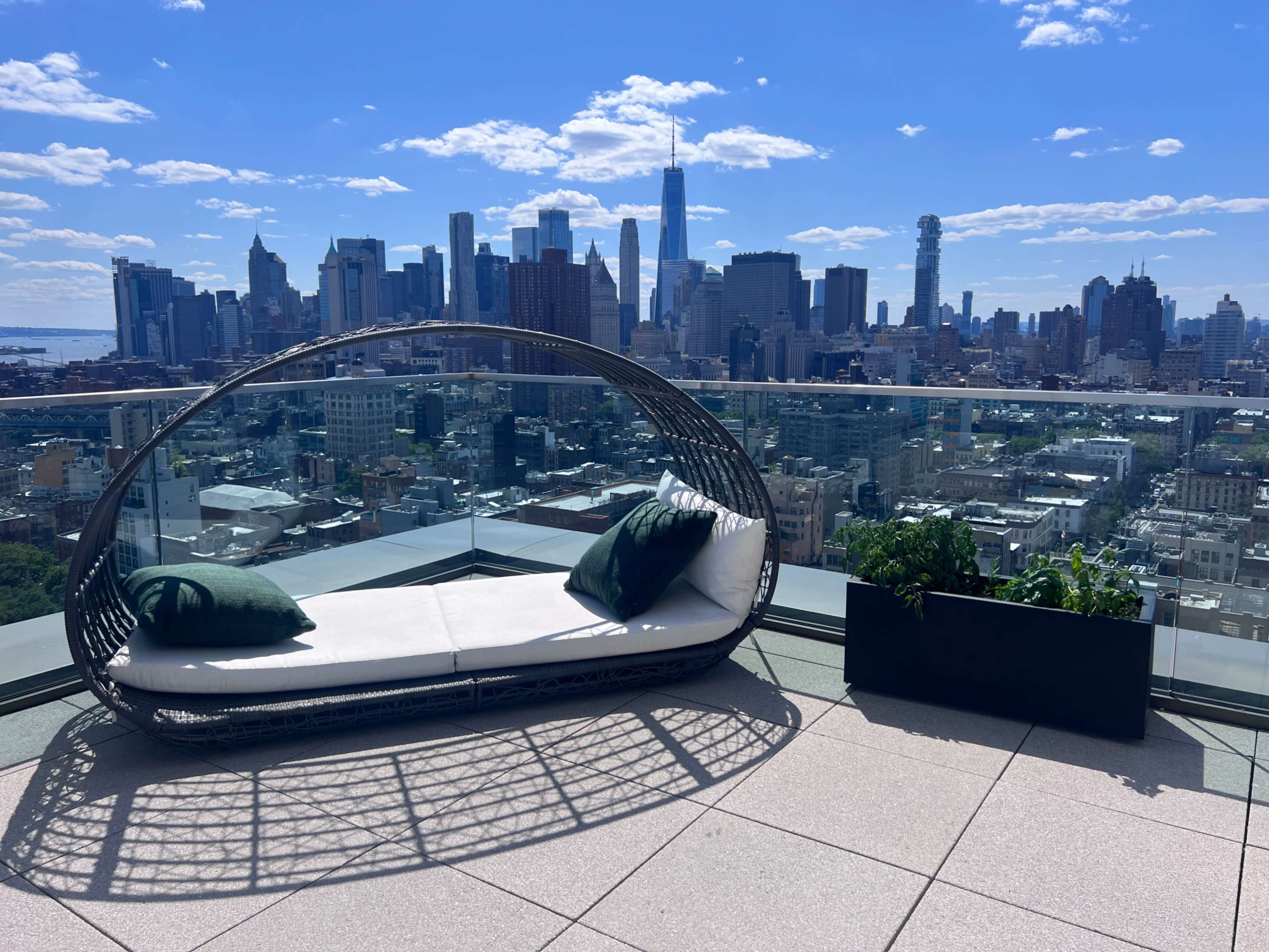 A modern outdoor seating area with a curved lounge chair and potted plants, overlooking a city skyline under a clear blue sky.