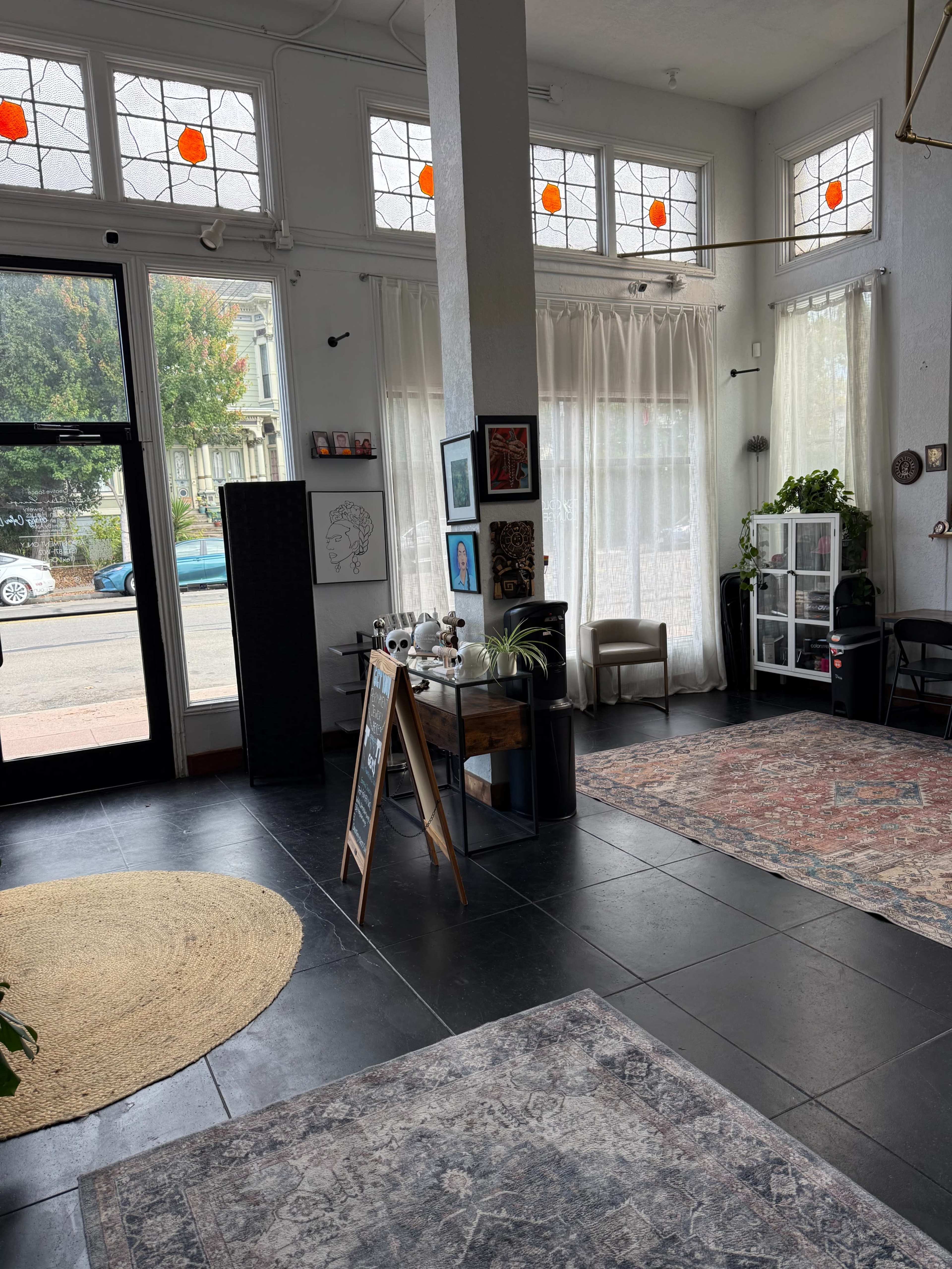The image shows a bright interior of a modern establishment featuring large windows, plants, and various decorative items placed around the space, including a rug and a signboard.