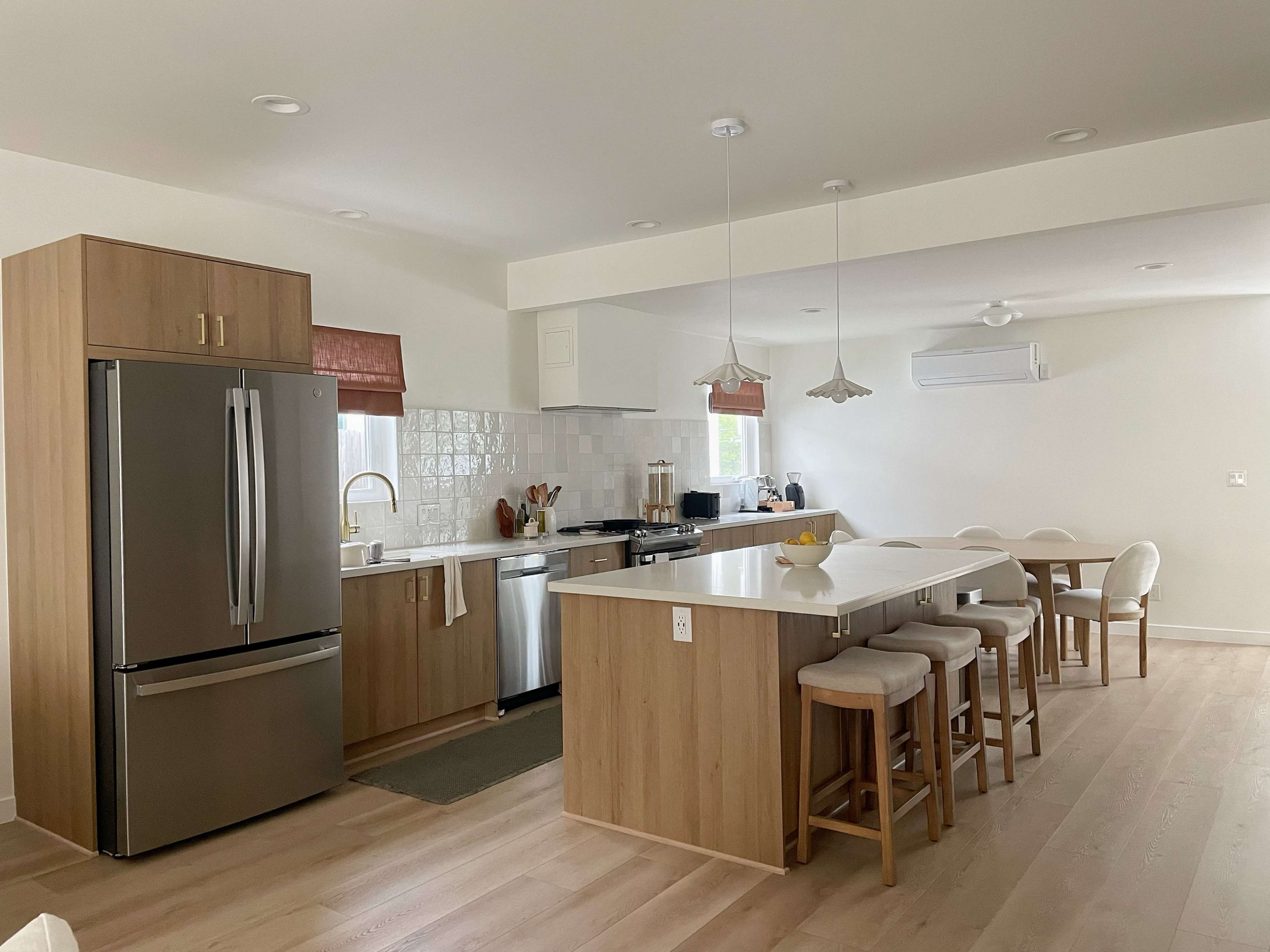 The image shows a modern kitchen featuring wooden cabinetry, stainless steel appliances, and a dining area with a round table and chairs.