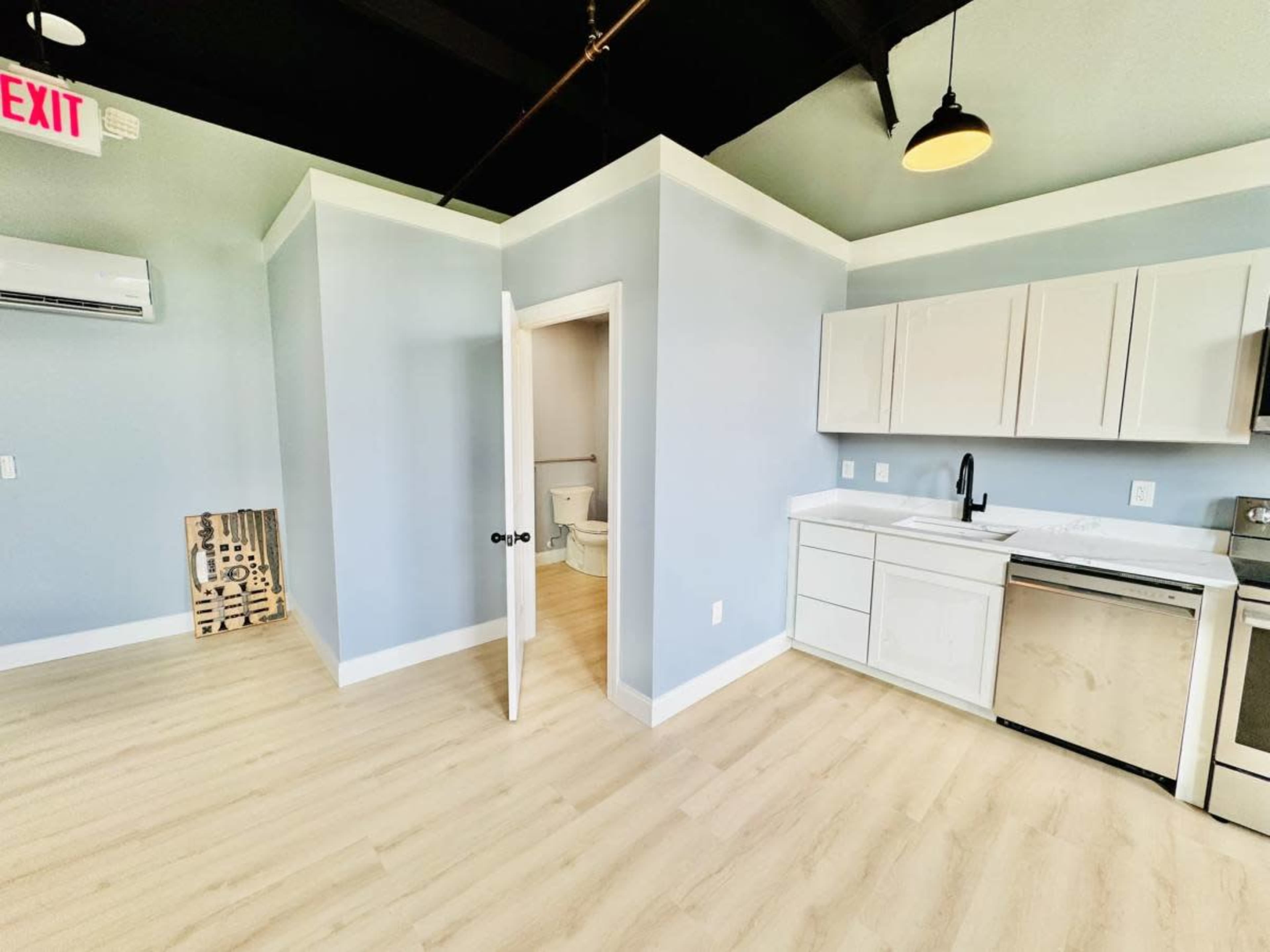 The image shows a small, modern open kitchen area with white cabinets, a stainless steel refrigerator, and a bathroom door partially open, revealing a toilet inside.