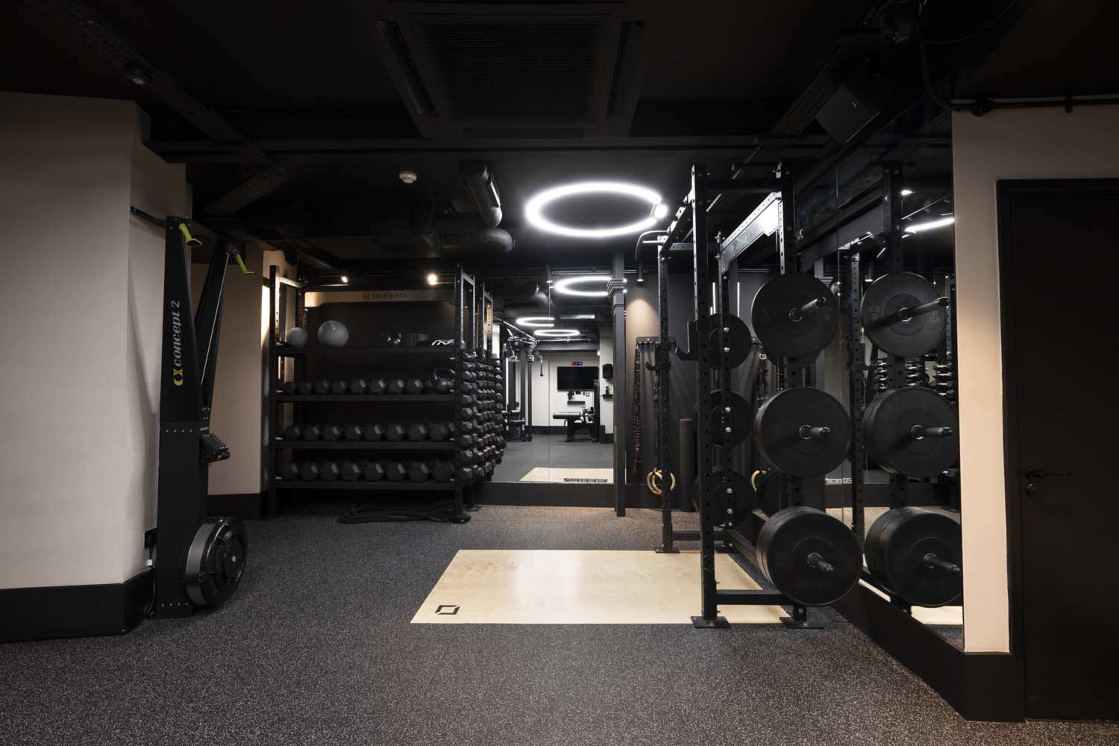 The image shows a modern gym interior featuring weightlifting equipment, free weights organized on racks, and a rubber flooring surface in a well-lit space.