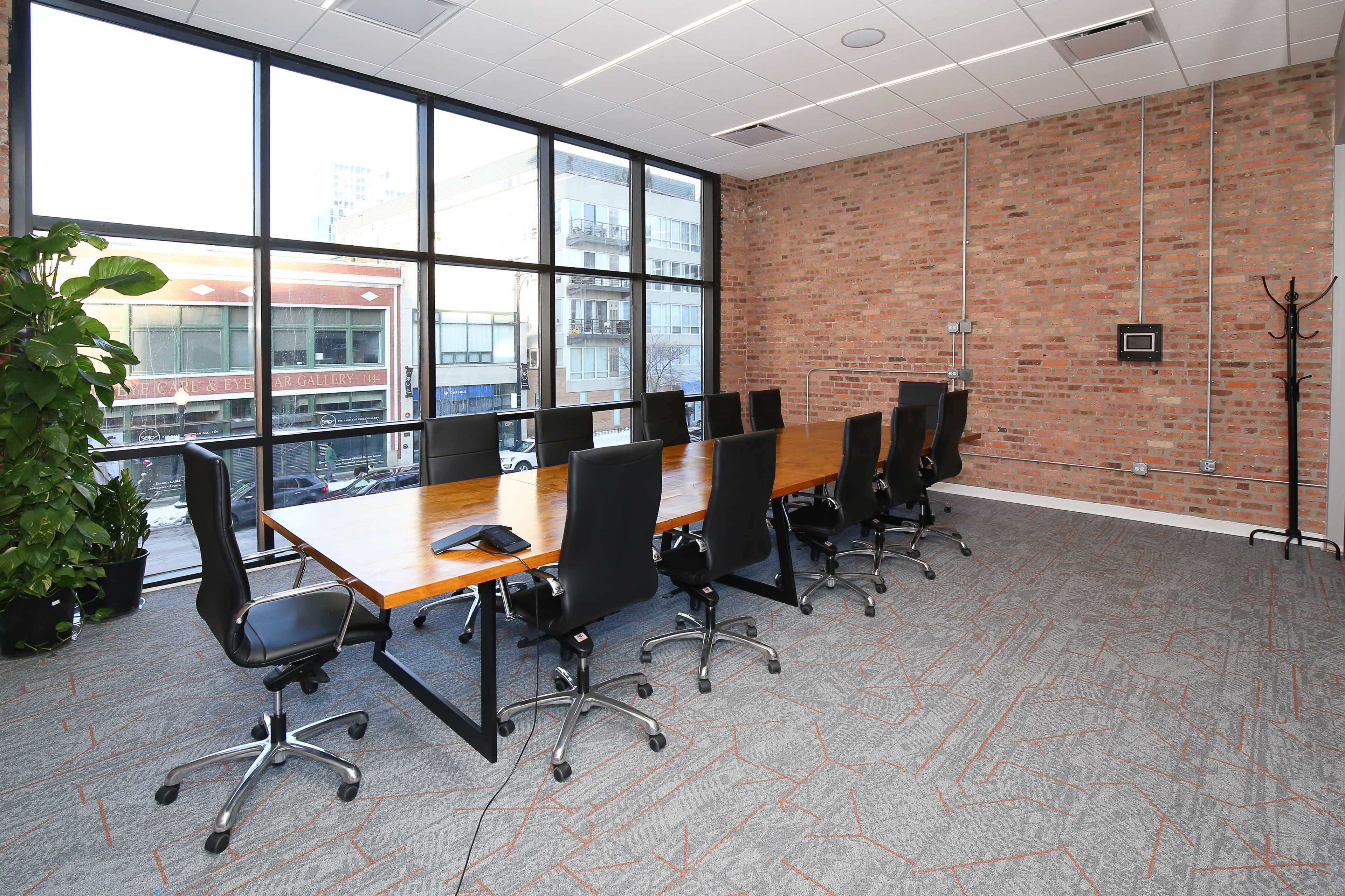 A modern conference room features a large wooden table surrounded by black rolling chairs, with large windows providing a view of the outside.