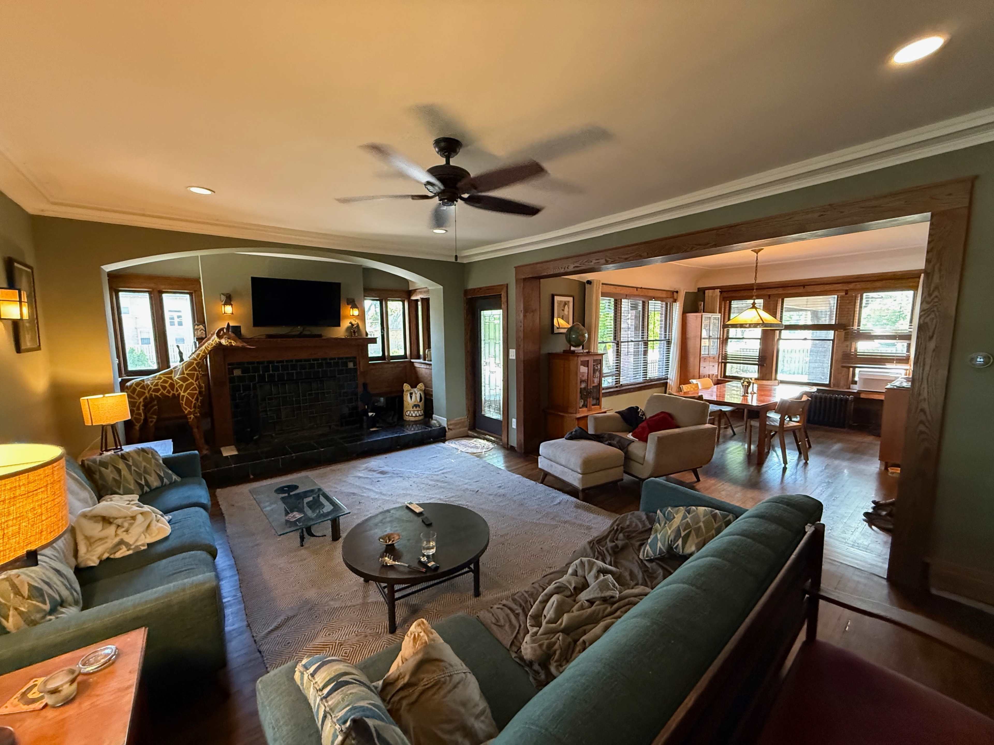 The image shows a spacious living room with a fireplace, a ceiling fan, and a view into an adjacent dining area, featuring various pieces of furniture and large windows.