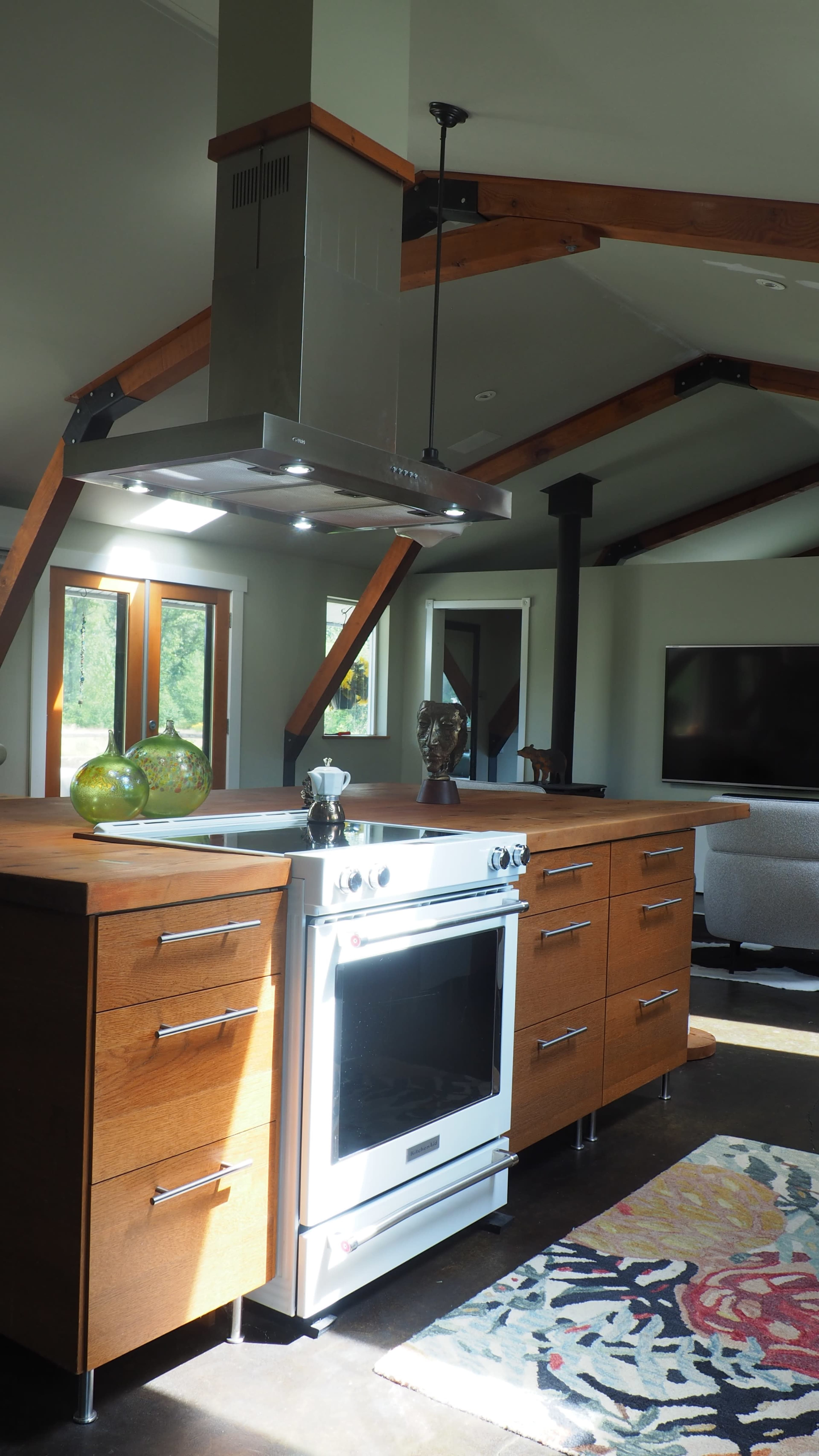 The image shows a modern kitchen with an island, wooden cabinetry, a stainless steel stove, and exposed beams in the ceiling.