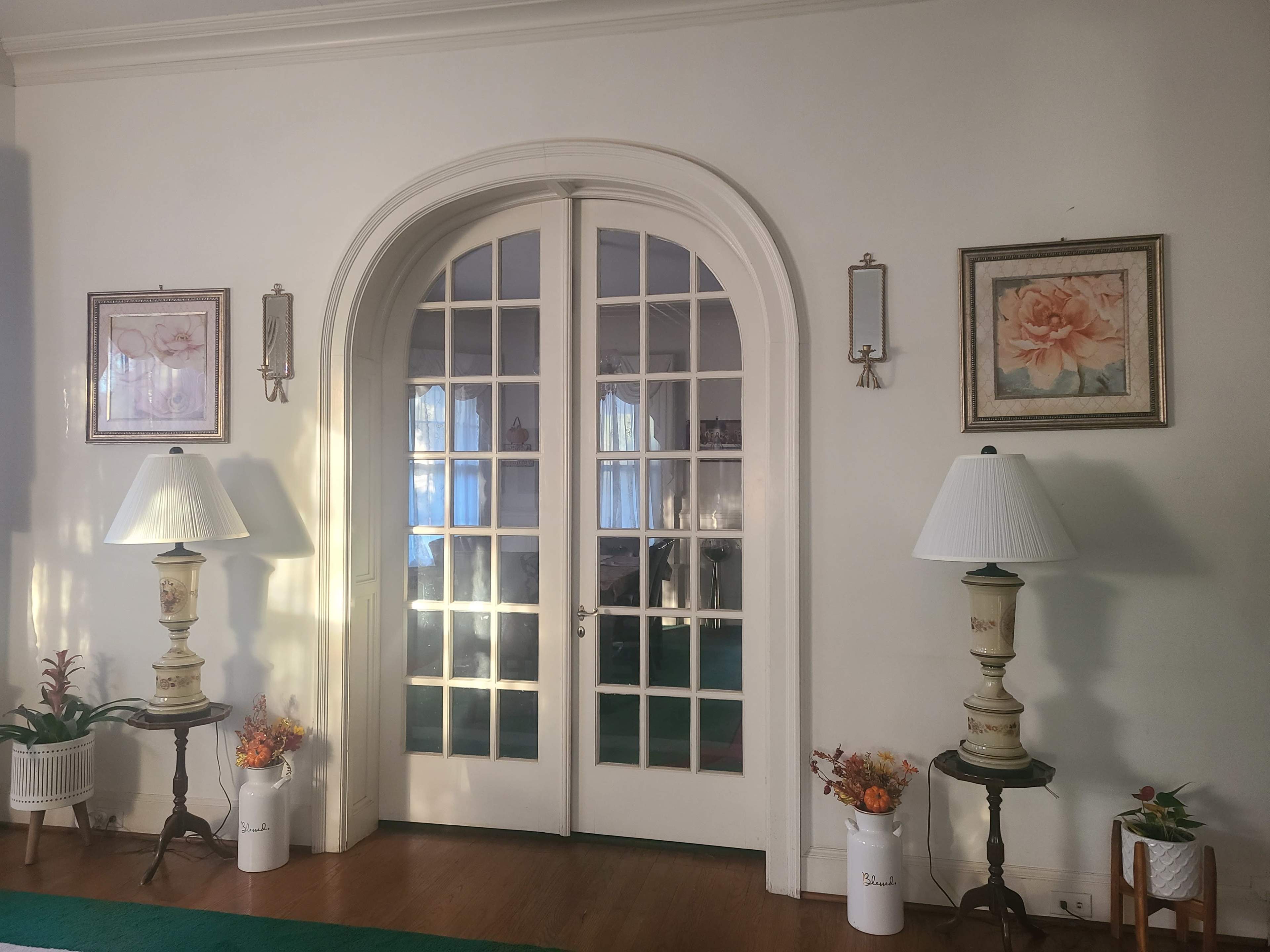 The image shows a room with a pair of French doors framed by two lamps on decorative tables, flanked by two floral paintings on the walls.