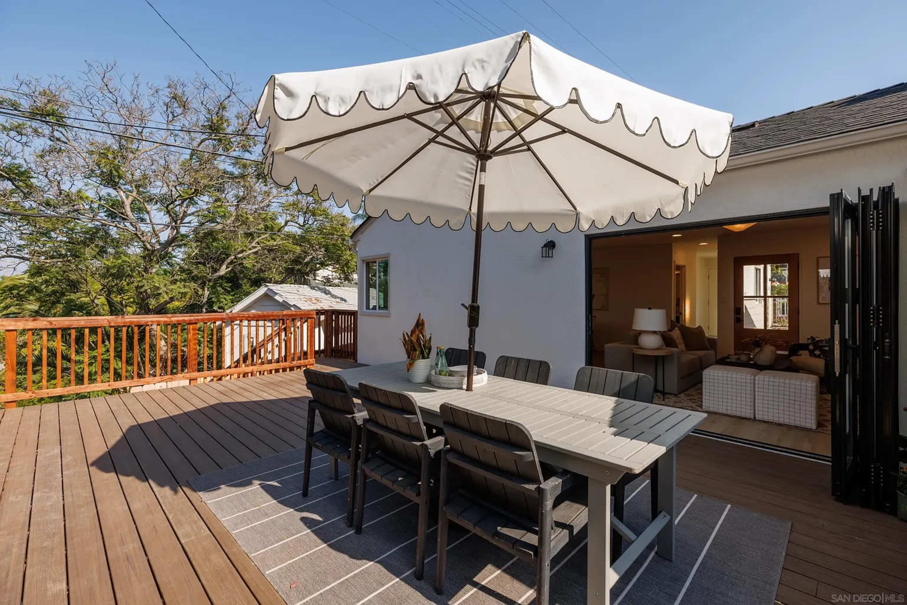 A spacious wooden deck features a large umbrella over a dining table with chairs, adjacent to an open door leading into a living area.