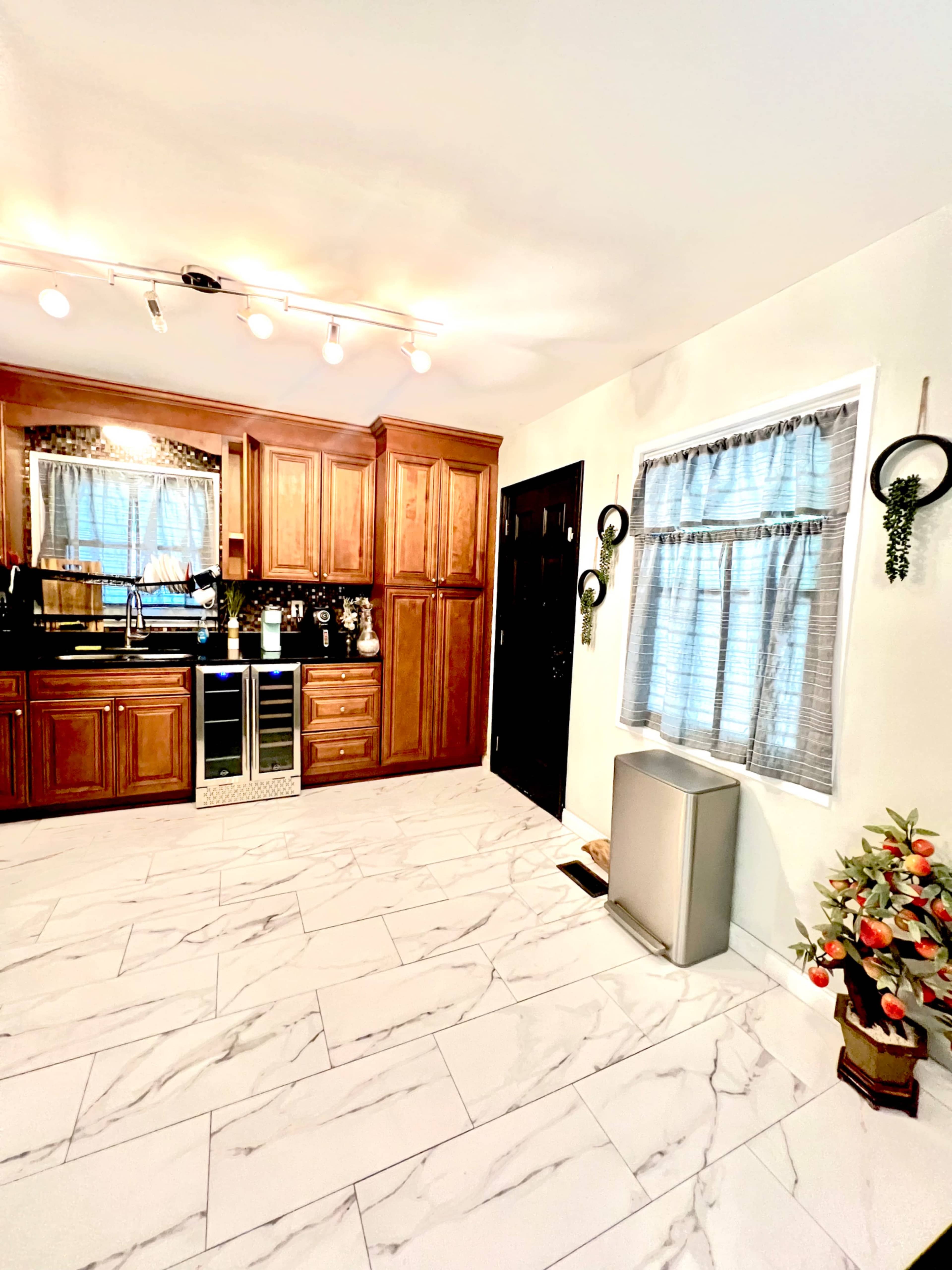 The image shows a kitchen with wooden cabinetry, a marble tile floor, a black front door, and a small window with sheer curtains, alongside a potted plant.